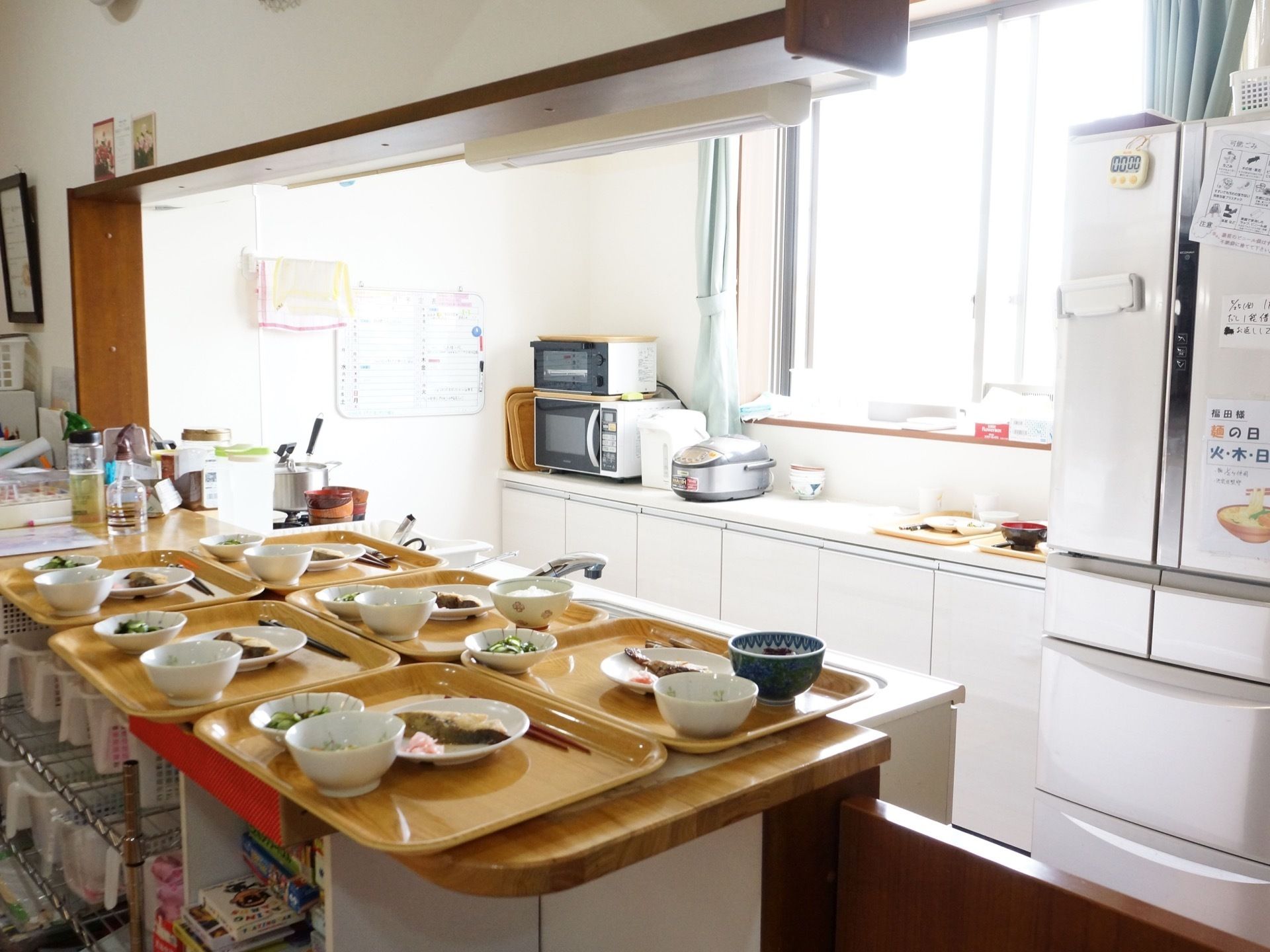 対面式カウンターキッチンである。窓側に冷蔵庫と炊飯器、電子レンジが置かれている。カウンター上には、お食事が配膳されている。