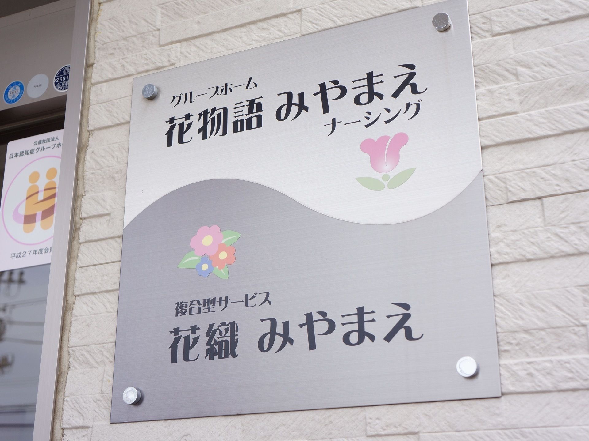 入り口脇の壁に掲げられているプレート。正方形のプレートにはグループホーム名と複合型サービス施設名が表示されている。