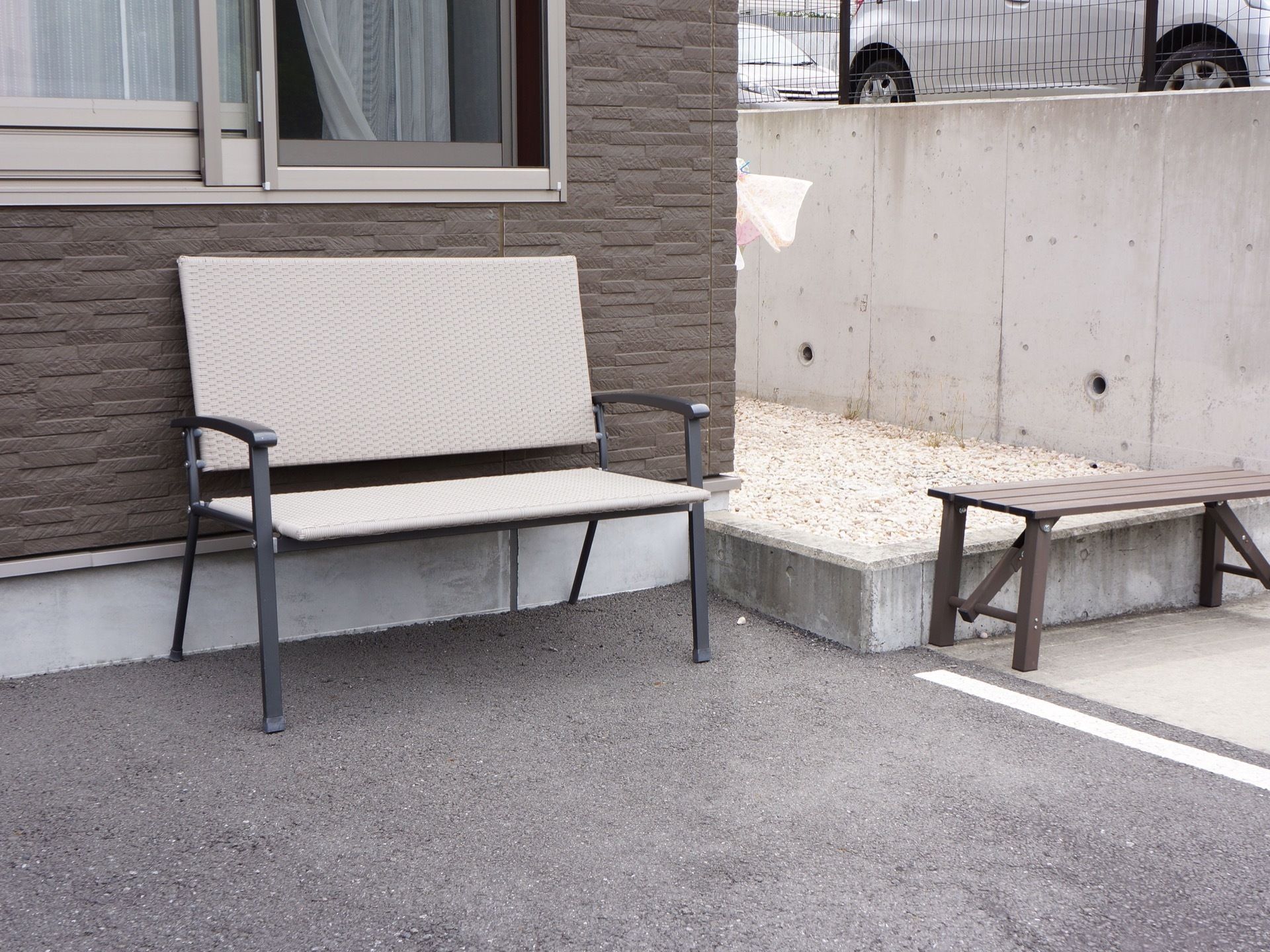 施設の建物の壁際に背もたれのある鉄製のベンチと長椅子の２脚が置かれている。入居者様が外出時に一休みするためのスペースとしてご利用いただける。
