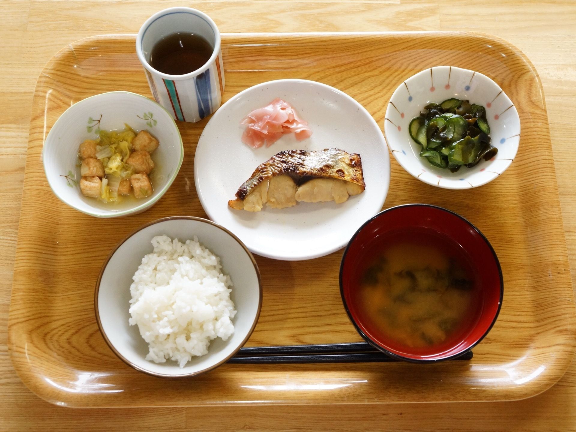 中央にメインの焼き魚と両脇にきゅうりの酢の物などの副菜が２皿と味噌汁に白いご飯、お茶がトレイに並んでいる。