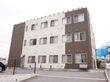 広い駐車場からの眺め。白と茶色の三階建ての四角い建物がある。窓が並んでおり、部屋数がたくさんあることが伺える。