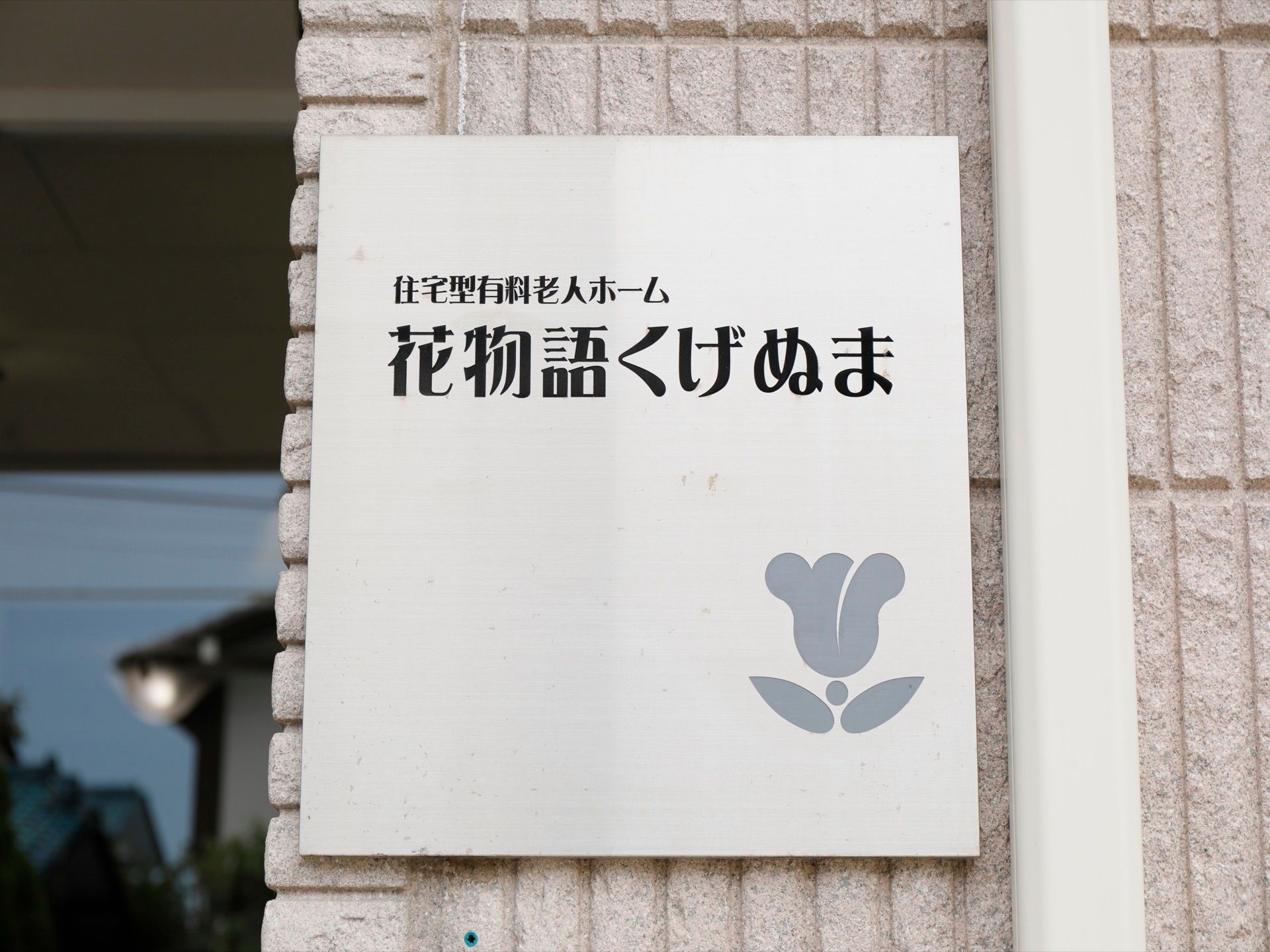 エアコン室外機のパイプの横にある施設看板。正方形のボードの中に施設名のロゴと花をかたどったマークが入っている。