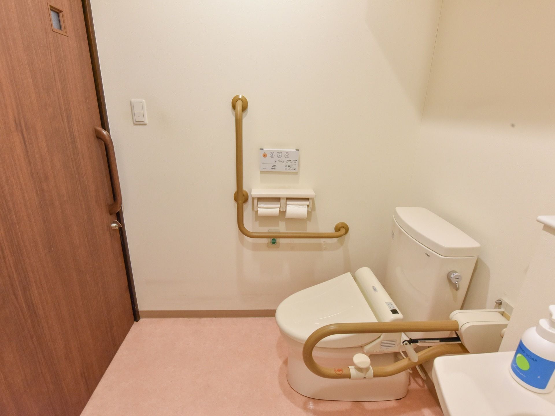木目調のスライド式のドアの正面に便座を備えたトイレである。エル型と可動式の手すりを設置し、横側に洗面台を設置している。