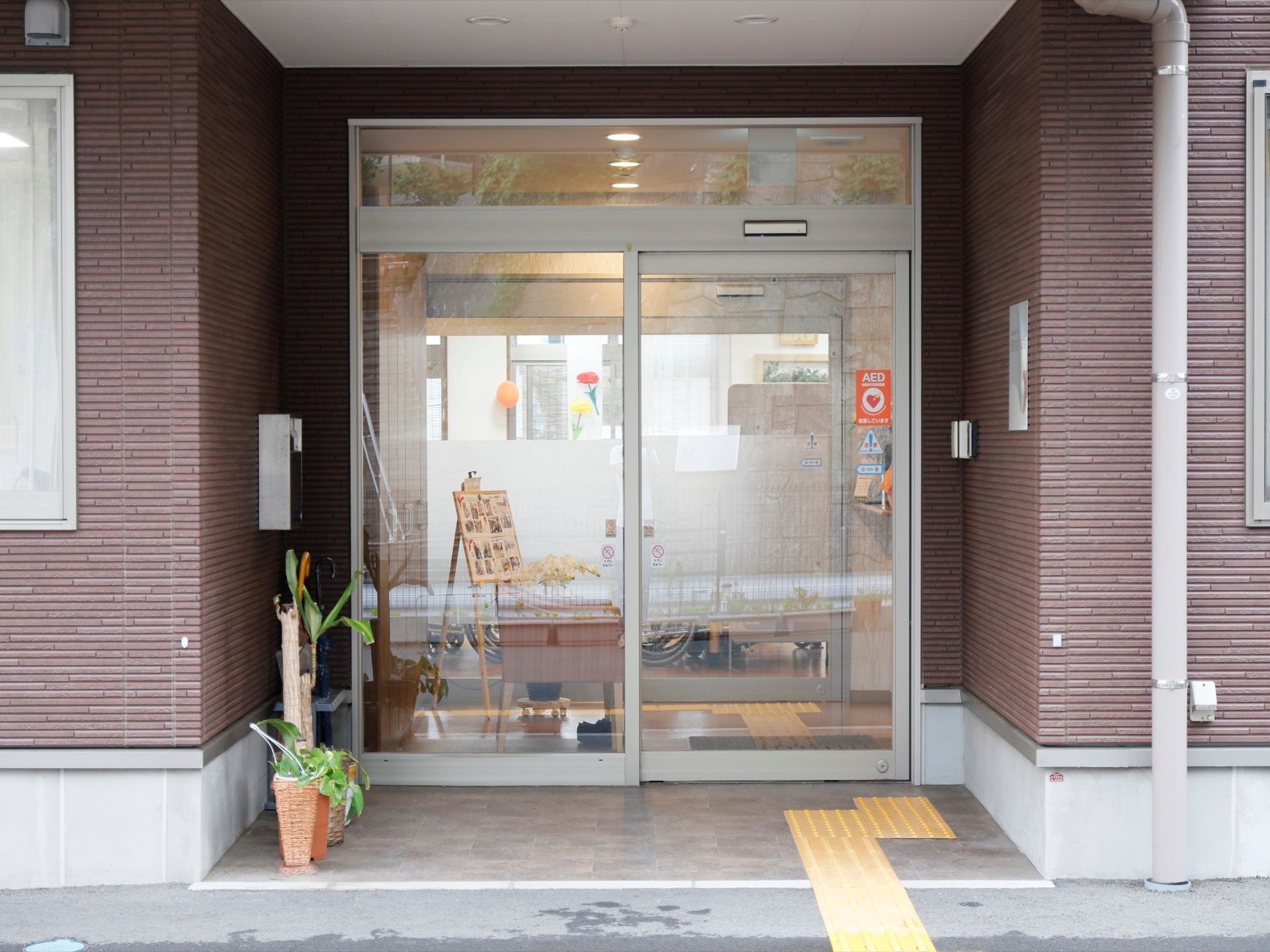 建物の外壁は、ブラウン系の落ち着いた色合いになっている。左端には傘立てがあり、その横に観葉植物が置いてある。