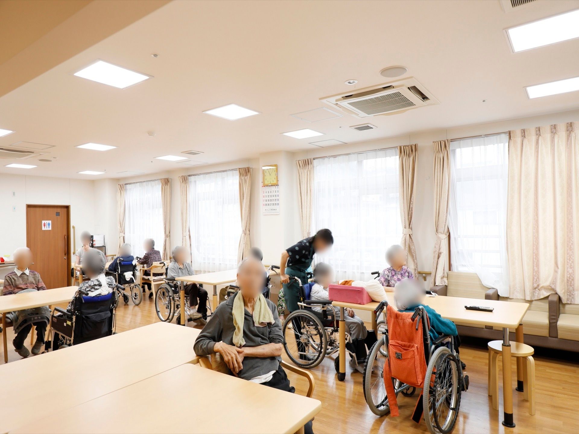 木目調デザインのテーブルの周りに、数多くの入居者様が座ってくつろいでいる。天井には、四角いライトが取り付けられている。