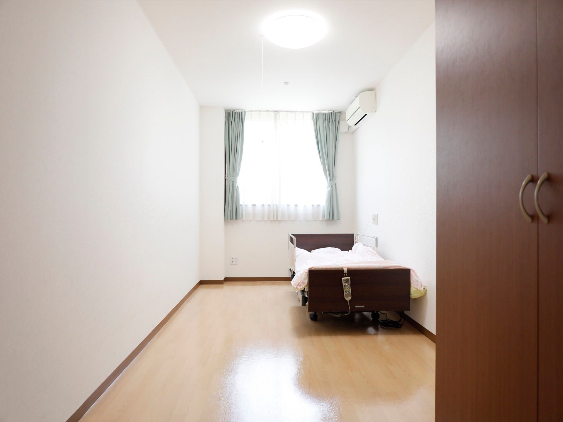 白色の壁に、グリーンのカーテンの付いた爽やかな印象の部屋である。介護ベッドやエアコン、クローゼットがある。