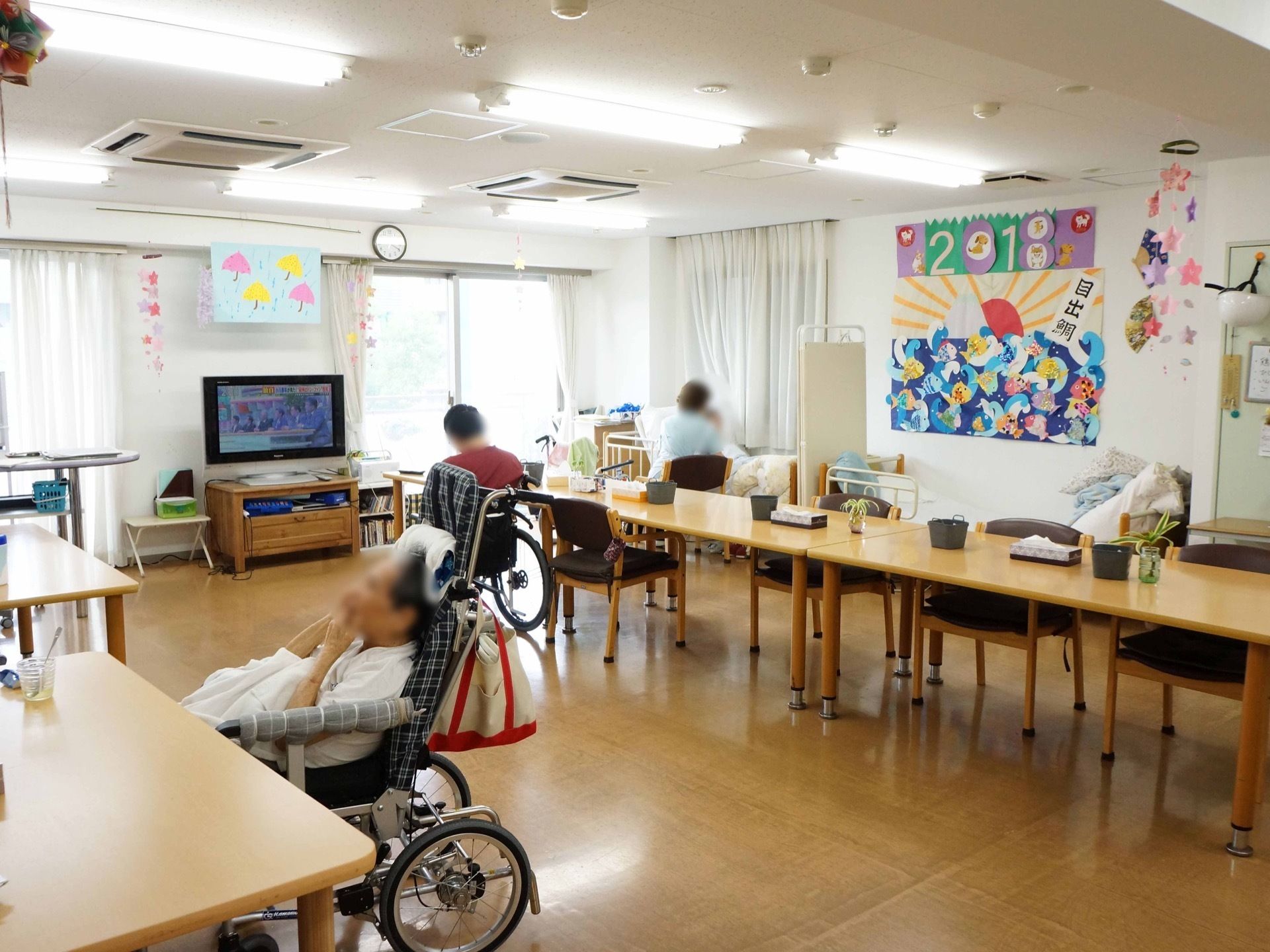 大型テレビが一台設置されている。奥には介護用ベッドが置かれ、寝たきりの入居者様も同じ食堂でお食事を摂っている。