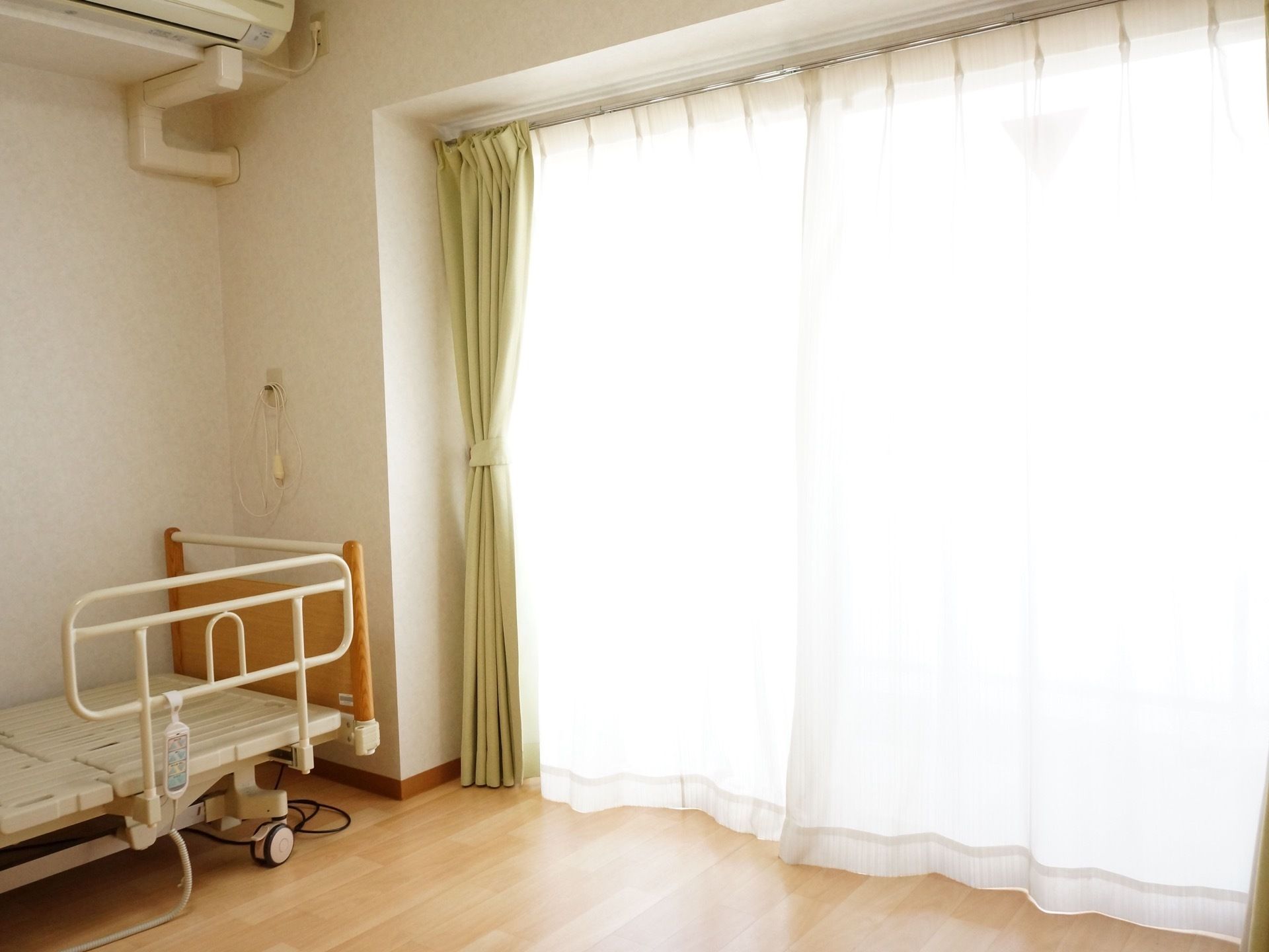 介護用ベッドが置かれている。その上にはエアコンが設置されている。枕元にはナースコールがある。窓は大きめのタイプである。