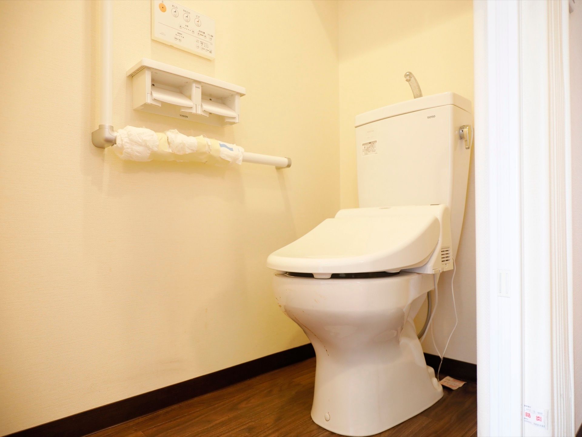 温水洗浄機能付きの洋式トイレである。壁にはリモコンやトイレットペーパーホルダーのほか、L字型の手すりも設置されている。
