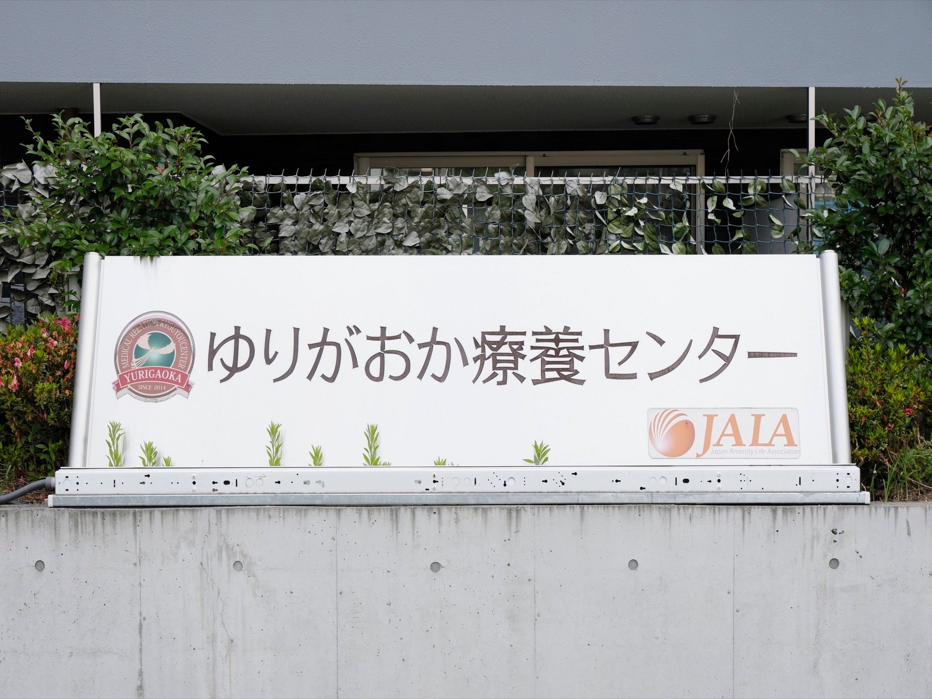 コンクリート塀の上に、大きな白地の看板がかかっている。大きな文字で、施設名と施設のマークなどが書かれている。