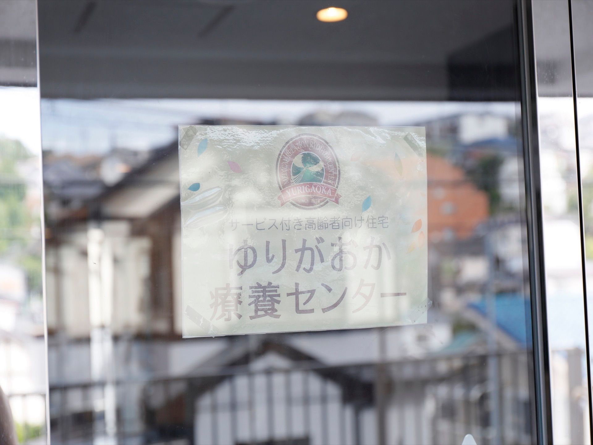 シルバーの枠のあるガラスのドアに、施設のロゴマークと名称がプリントされた紙が貼られている。紙はビニール袋に入っている。