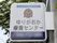 壁から正方形の看板が出ており、施設の名称が黒い文字で書かれている。その上にロゴマークがあり、看板の下部は紺色である。