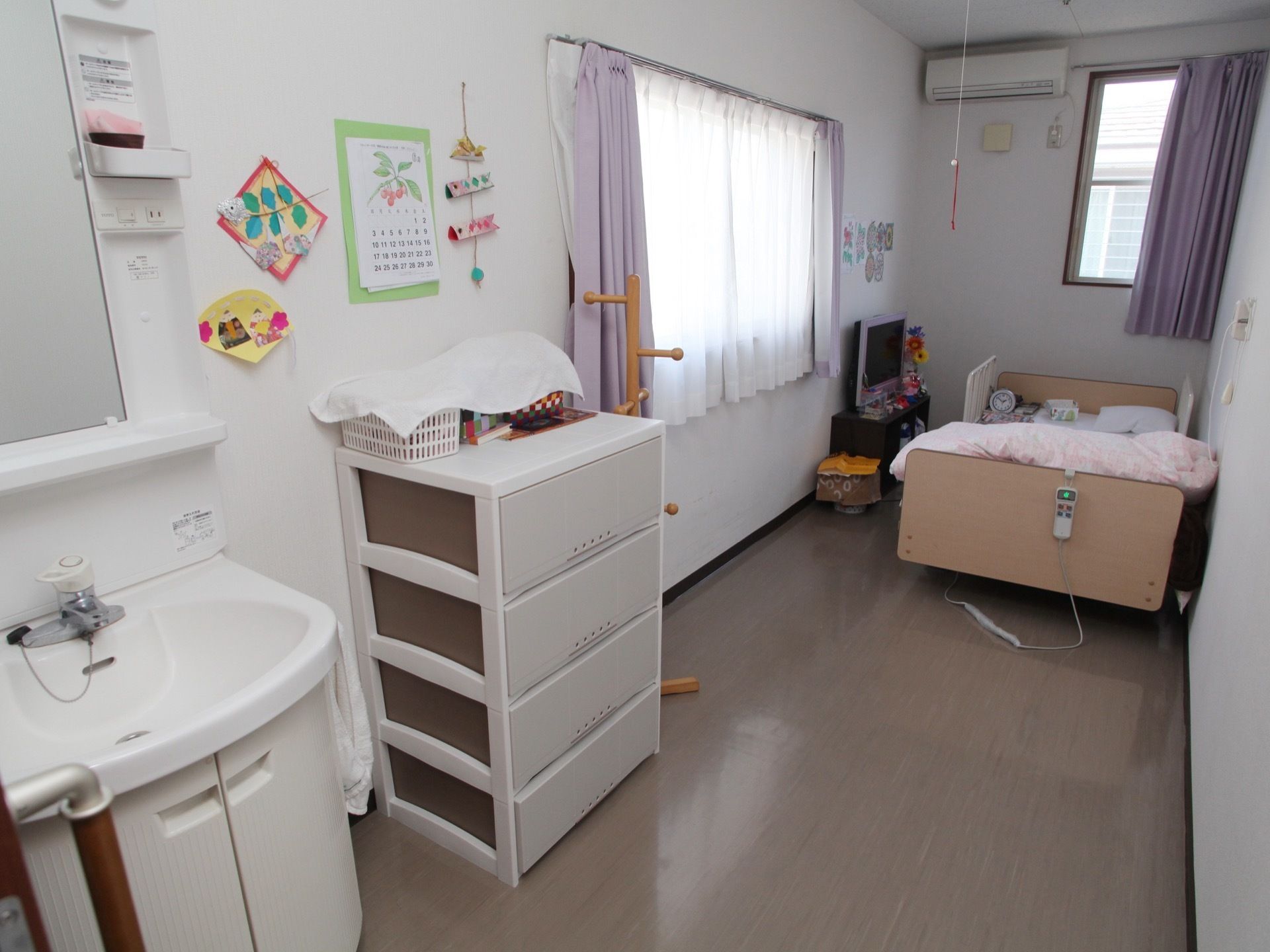 紫色のカーテンがアクセントになっている居室である。洗面台やチェスト、ベッドなどが完備され自宅のような雰囲気がただよう。