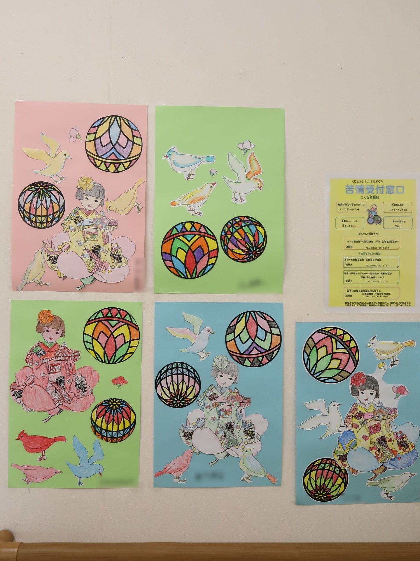 入居者様の塗り絵作品が壁に掲示されている。着物を着た女の子や手まりが丁寧に塗られ、入居者様の名前が書かれている。