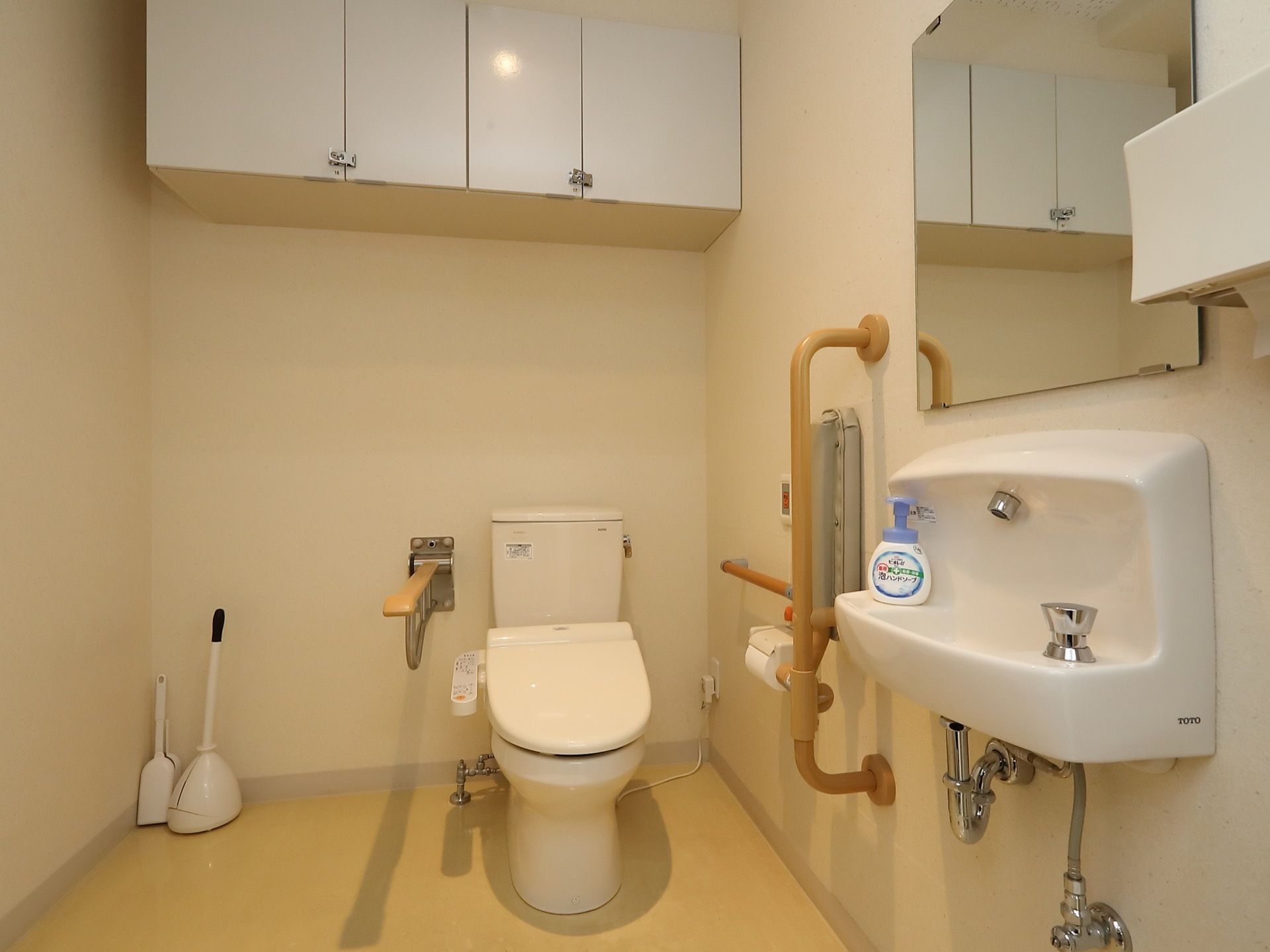 広いスペースにトイレが設置されている。便座の横に可動式の手すりがついている。壁据えつけ式の簡易洗面台がある。