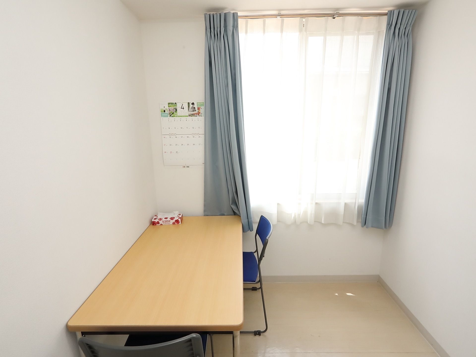 談話室の窓に、レースのカーテンと薄い青色のカーテンがかかっている。部屋の隅にテーブルが置かれている。