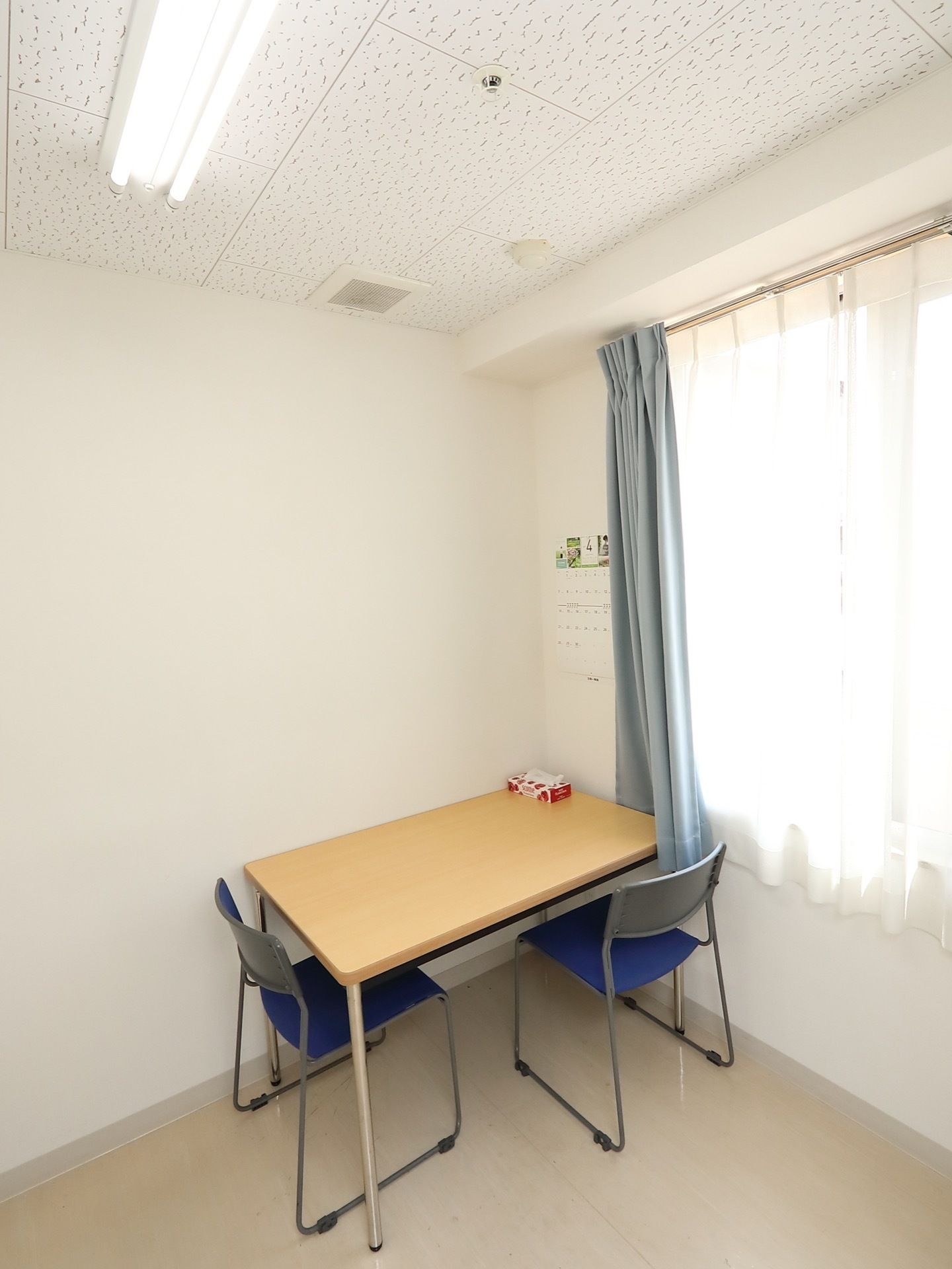 談話室の窓際に、壁につけられた状態でテーブルが置かれている。窓の横の壁には、月めくりのカレンダーがかかっている。
