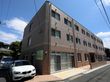 茶系統の色を基調とした３階建ての施設である。玄関前には低い壁が造られている。建物横に車が２台停まっている。