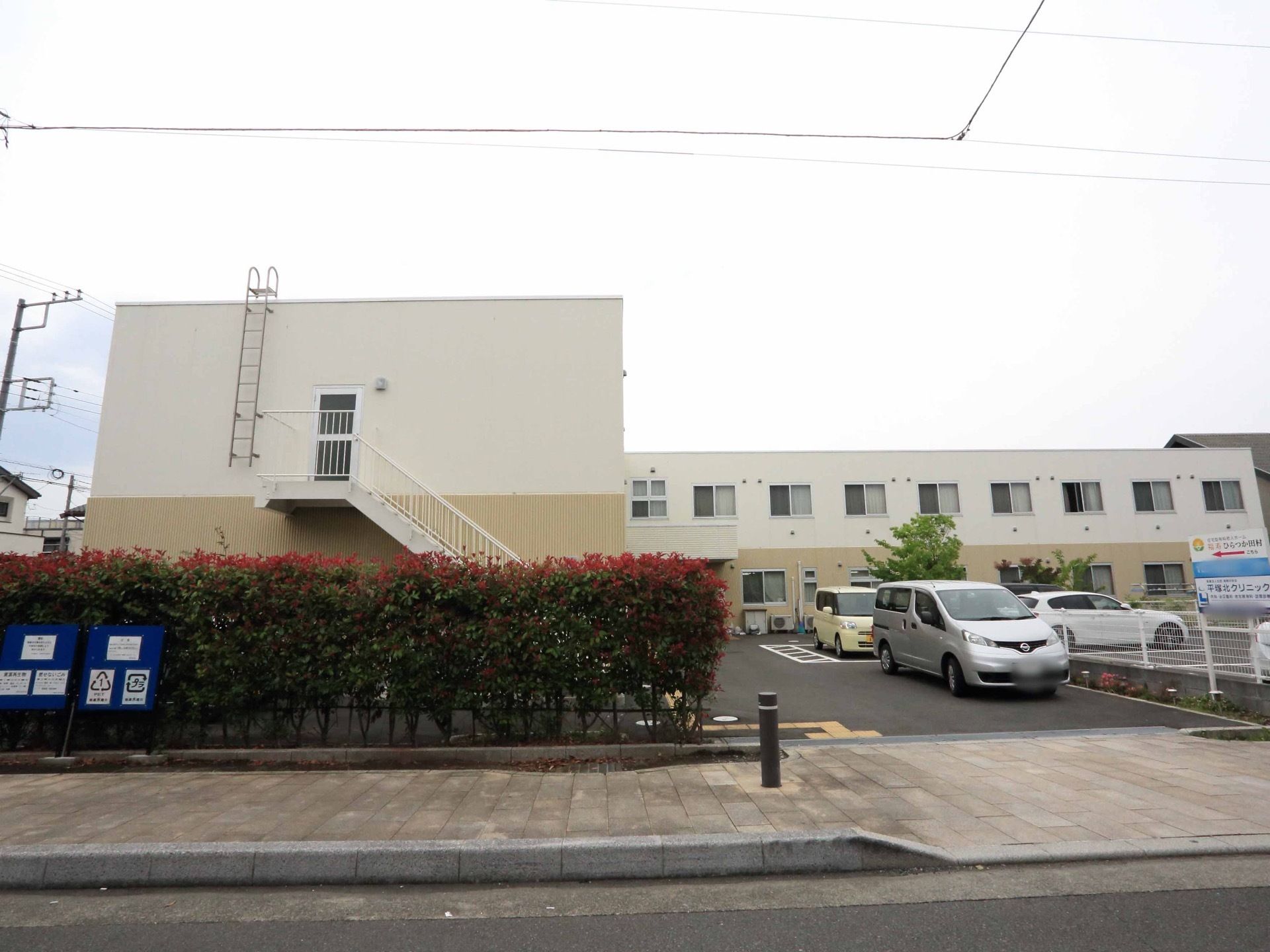 施設の建物の前には広々とした駐車場があり、数台の車が停まっている。駐車場の前は歩道になっていて、境に生垣がある。
