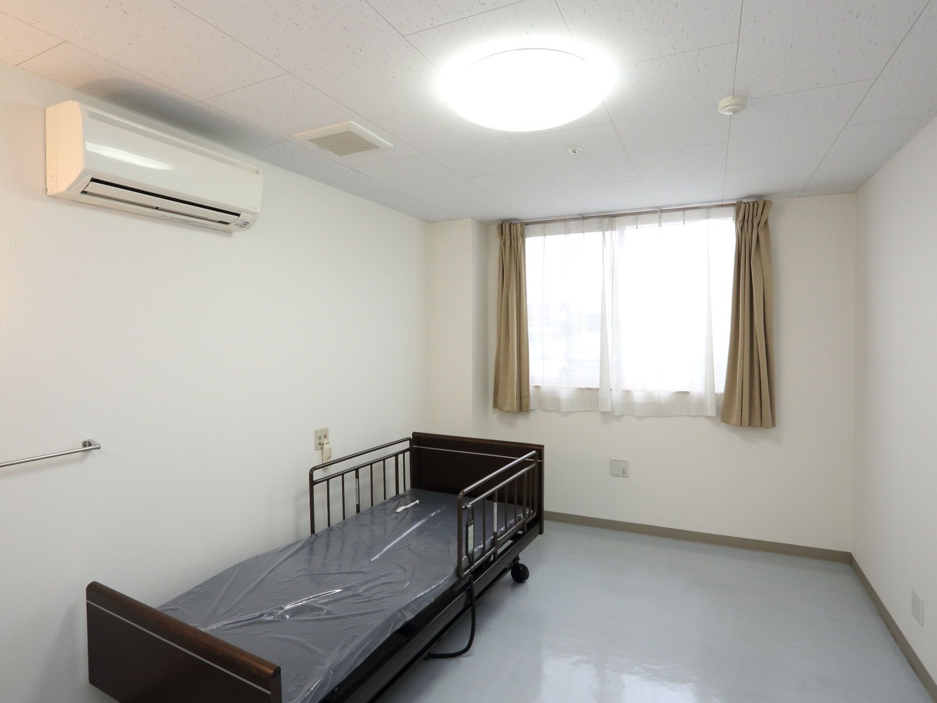 カーテン付きの窓のある居室には電動介護ベッドが置かれ、壁にはエアコン、ナースコールが取り付けられている。