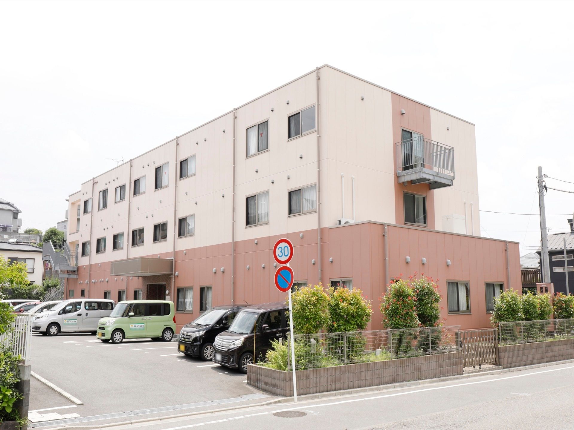 茶色とクリーム色の外壁の三階建てマンションタイプの外観で、施設の前には駐車場が完備されている。施設の奥には非常階段も見える。