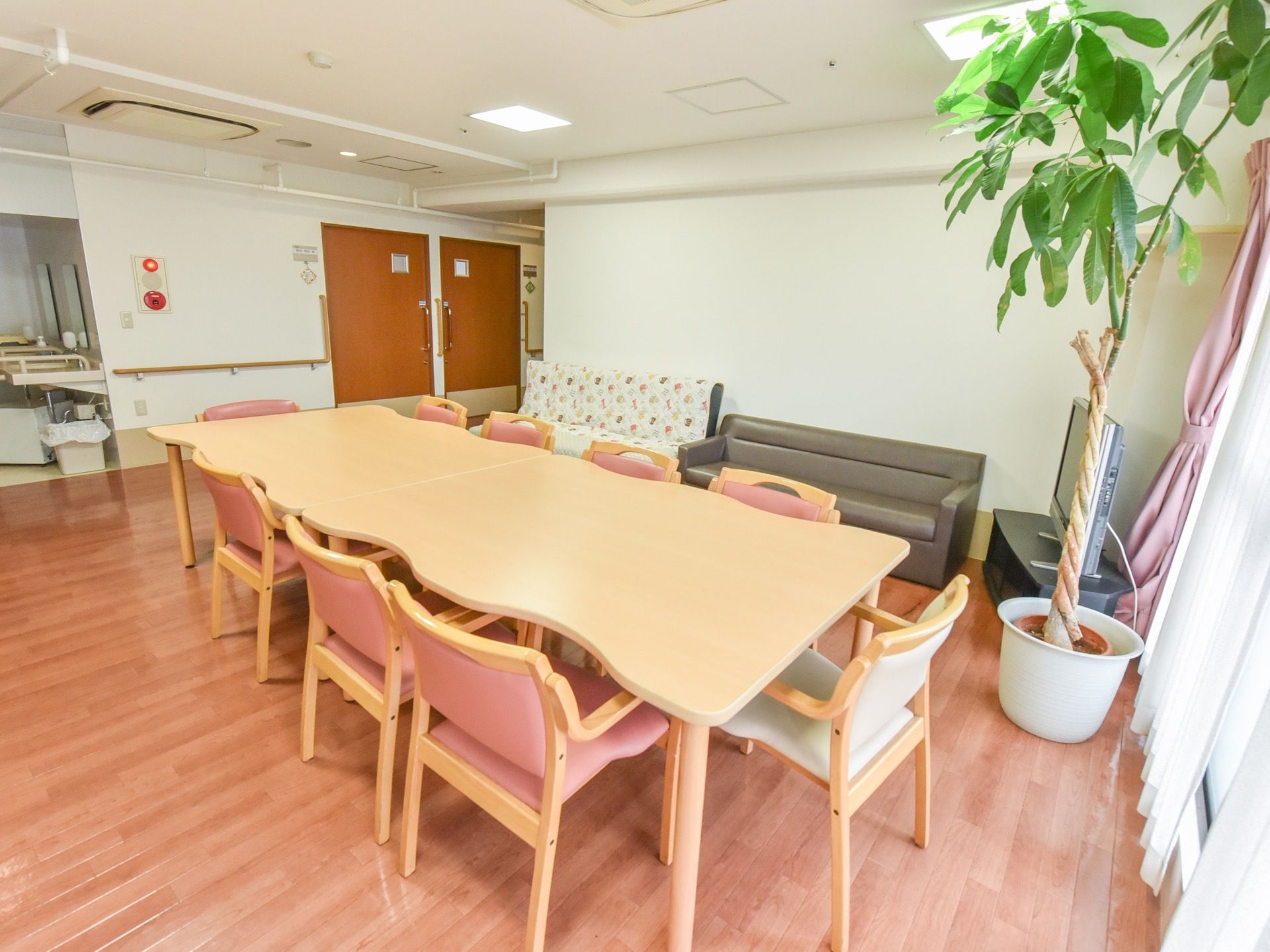 大きなテーブルとピンクの椅子が置かれた食堂。壁際にはソファーが２つと、大型のテレビが備え付けられている。