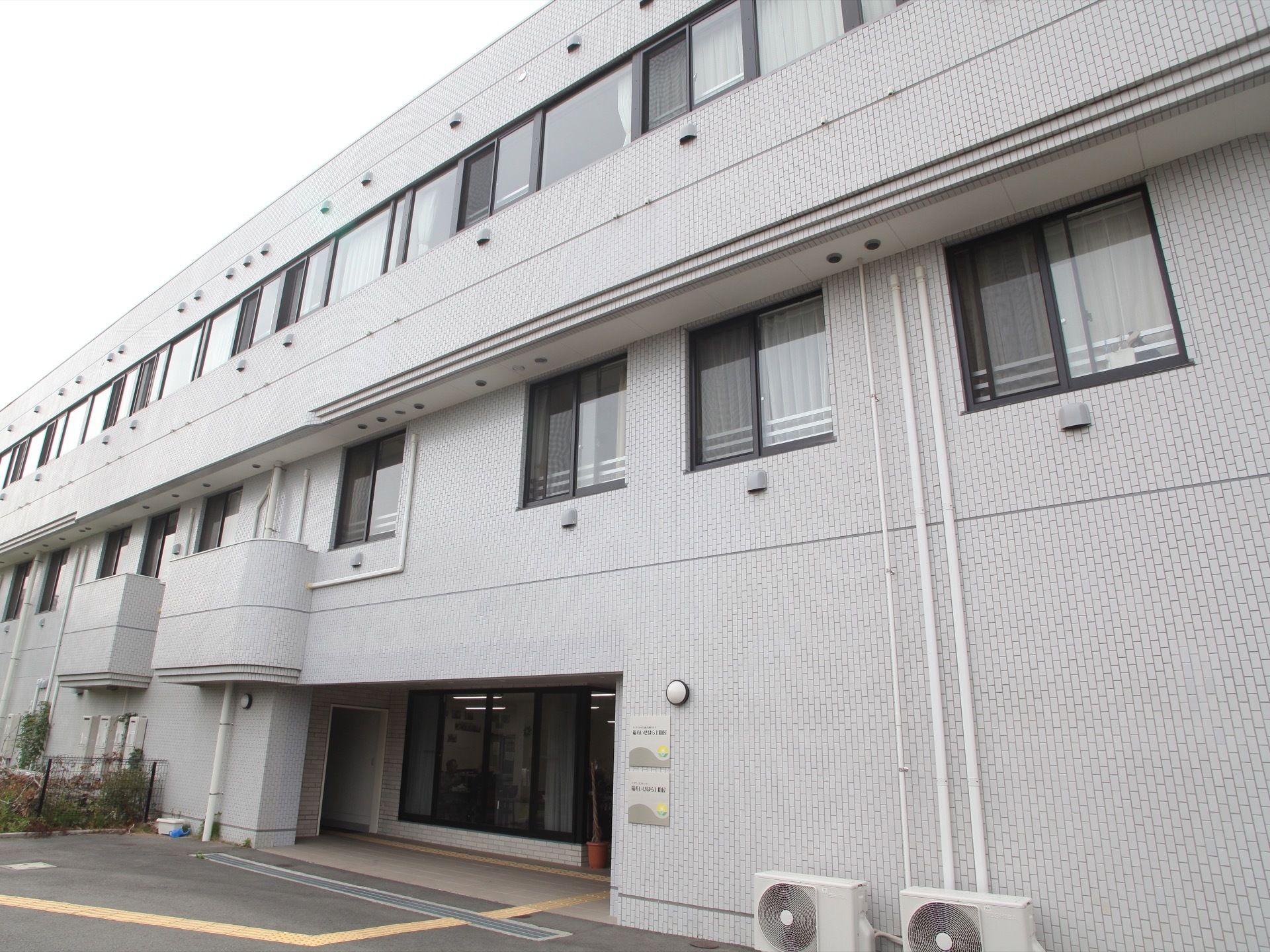 大きな白い建物で、窓がたくさんついている。壁際にはエアコンの室外機が置かれ、施設名の書かれたプレートが掲げられている。