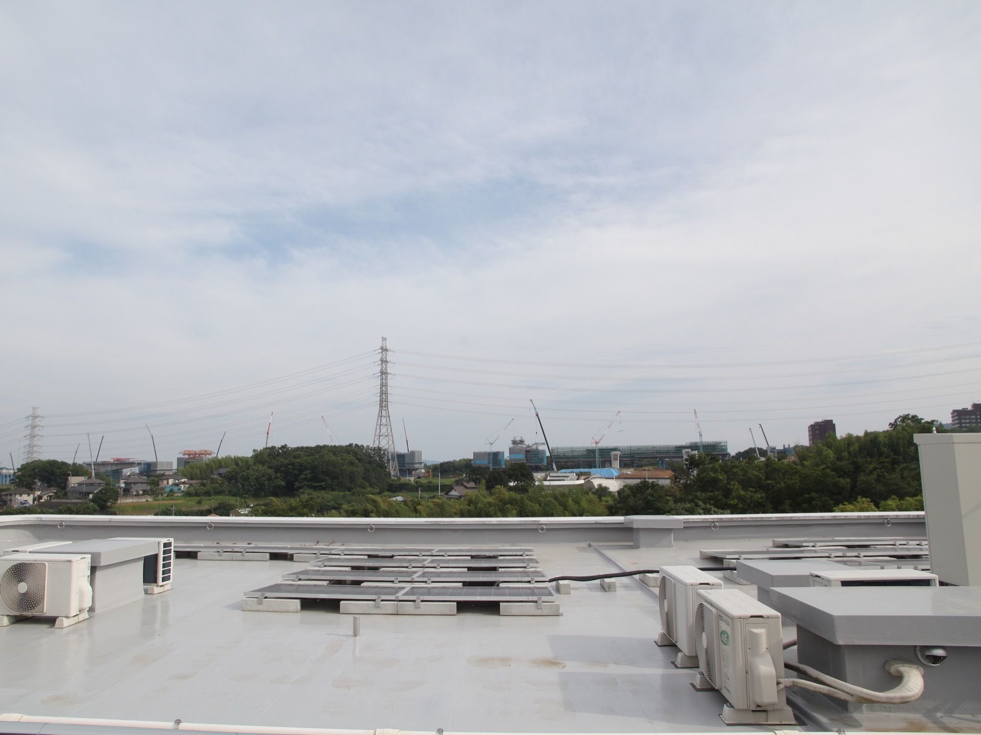 屋上にはエアコンの室外機や、太陽光発電のパネルが置かれている。施設のまわりには緑が多く、鉄塔が建っている。