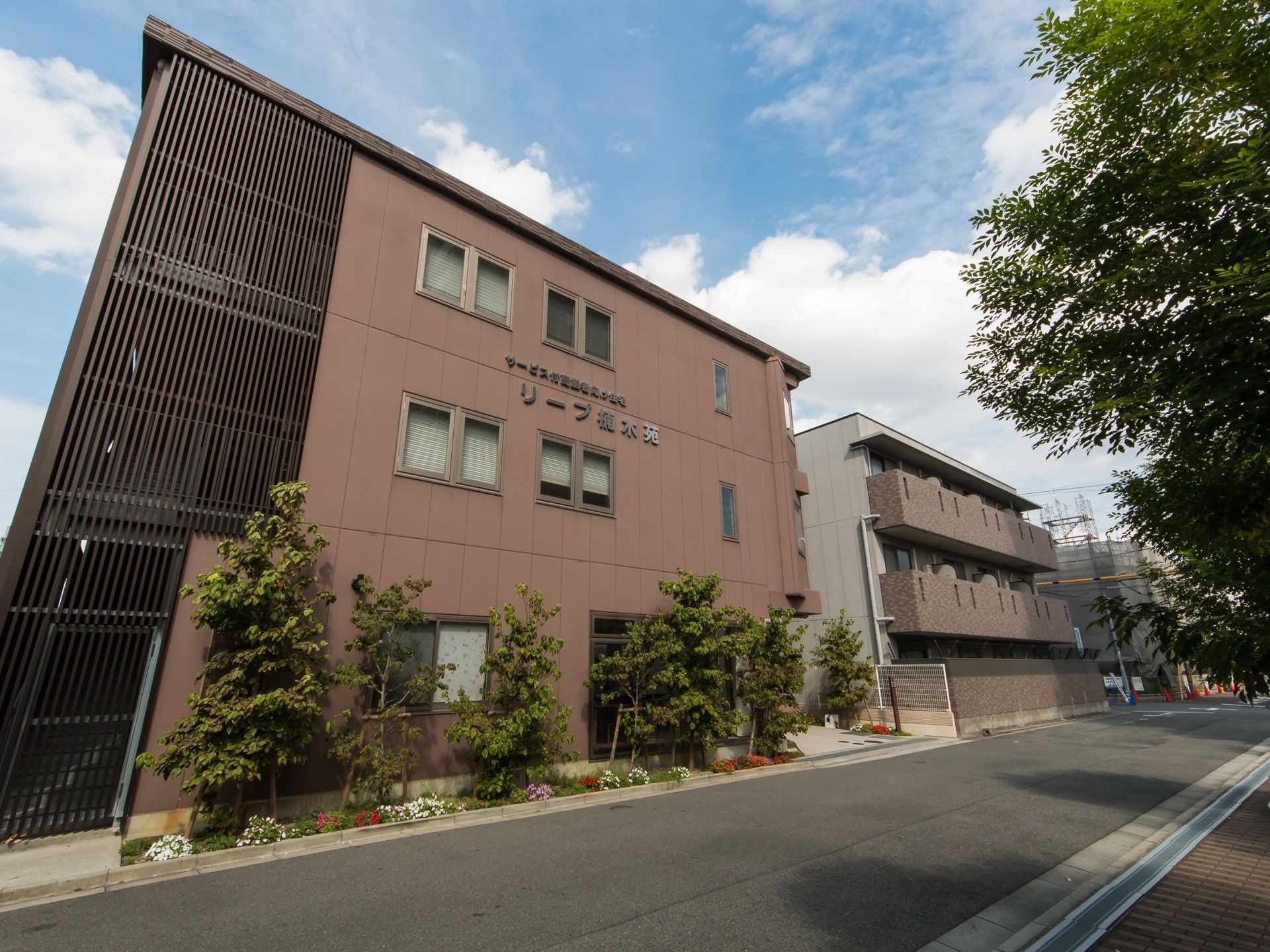 道路に面して建っている３階建ての施設である。道路をはさんだ向かい側には背の高い木が生えていて、道路に影を作っている。
