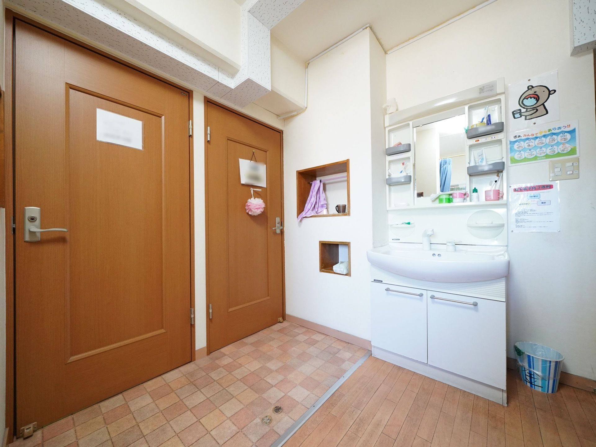 鏡と収納棚がついている洗面台がある。すぐ横にはドアが２つ並んでいる。壁には衛生マナーのポスターが貼られている。