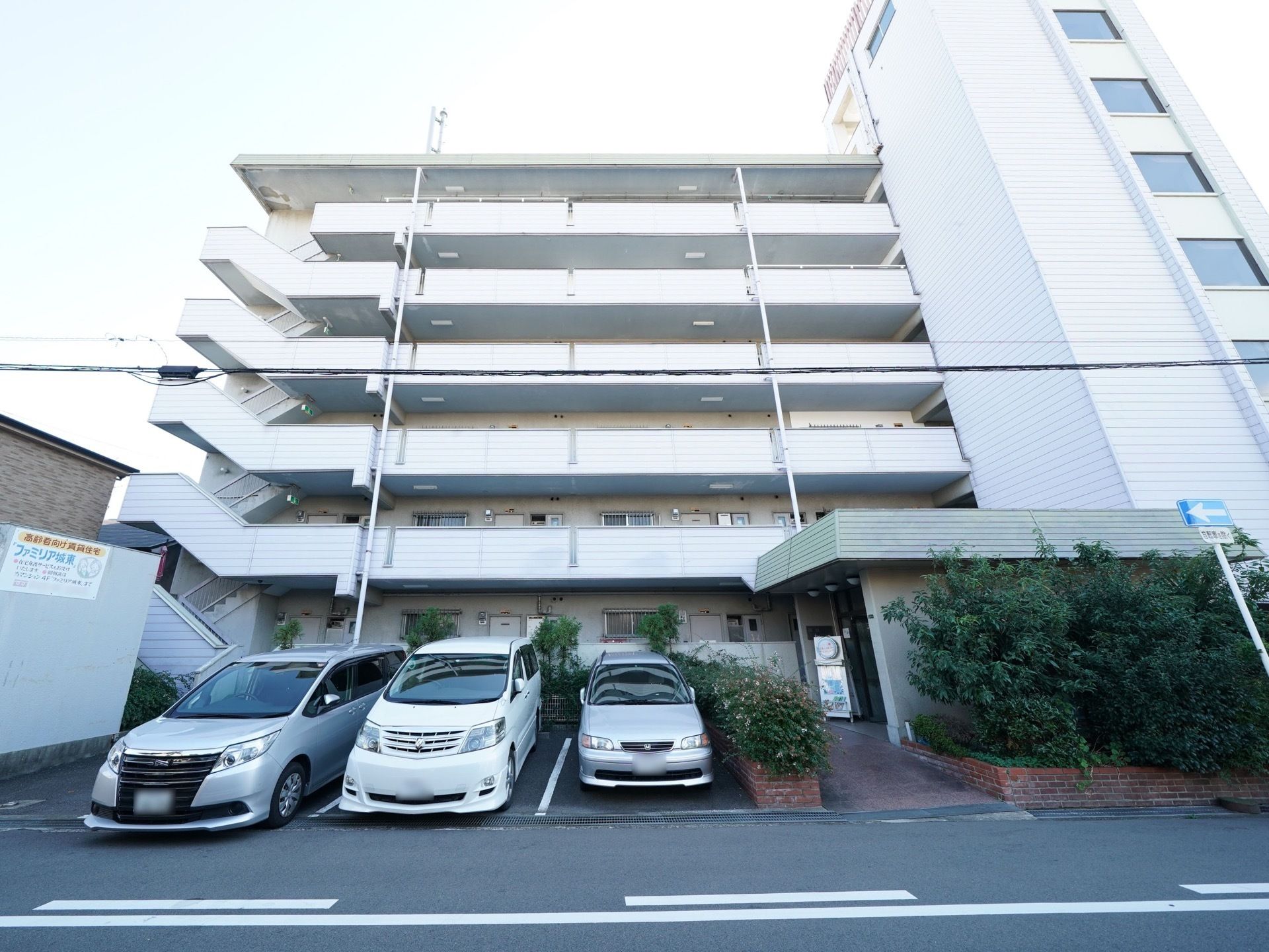 白い大きな鉄骨造りの建物。目の前は駐車場になっており、玄関先には植え込みがある。外つけの非常階段がある。