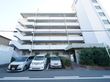 白い大きな鉄骨造りの建物。目の前は駐車場になっており、玄関先には植え込みがある。外つけの非常階段がある。