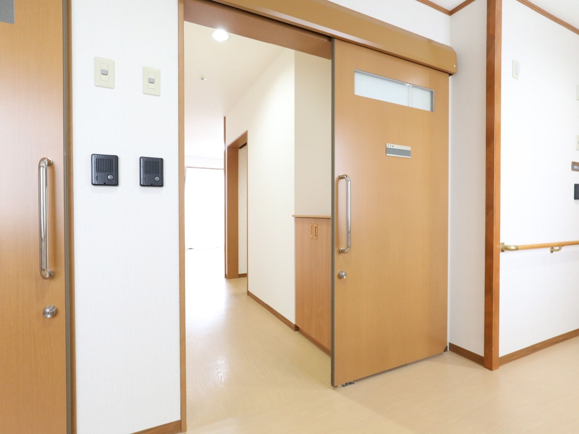 通路側から居室のスライドドア、さらに部屋の奥を見た様子である。内部は大きな窓から光が射し込み明るい。入り口にインターホンが付いている。