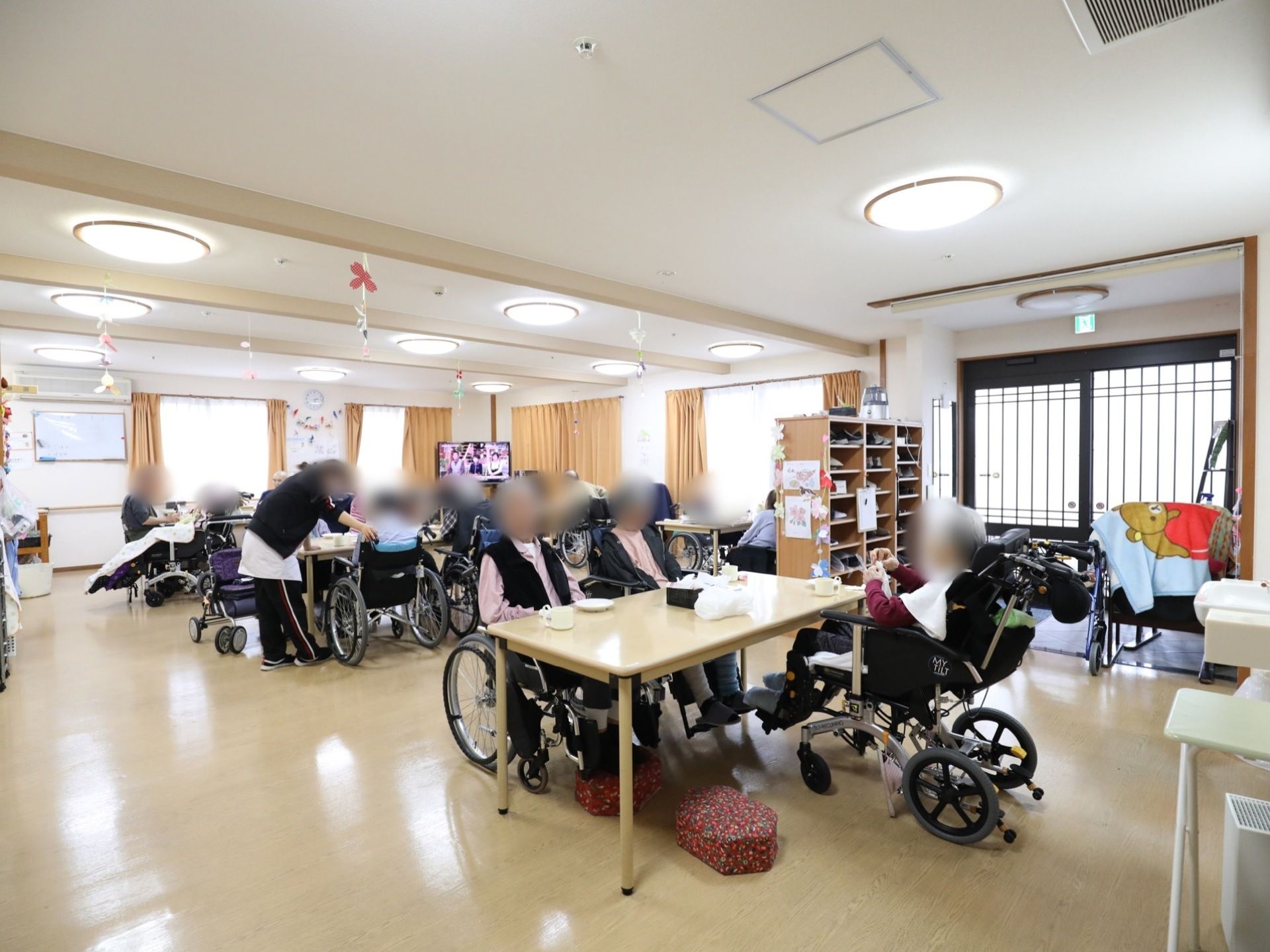 窓からの光や複数の照明器具により大変明るい共用スペースに複数のテーブルが並び、車椅子を利用する入居者が集まっている。