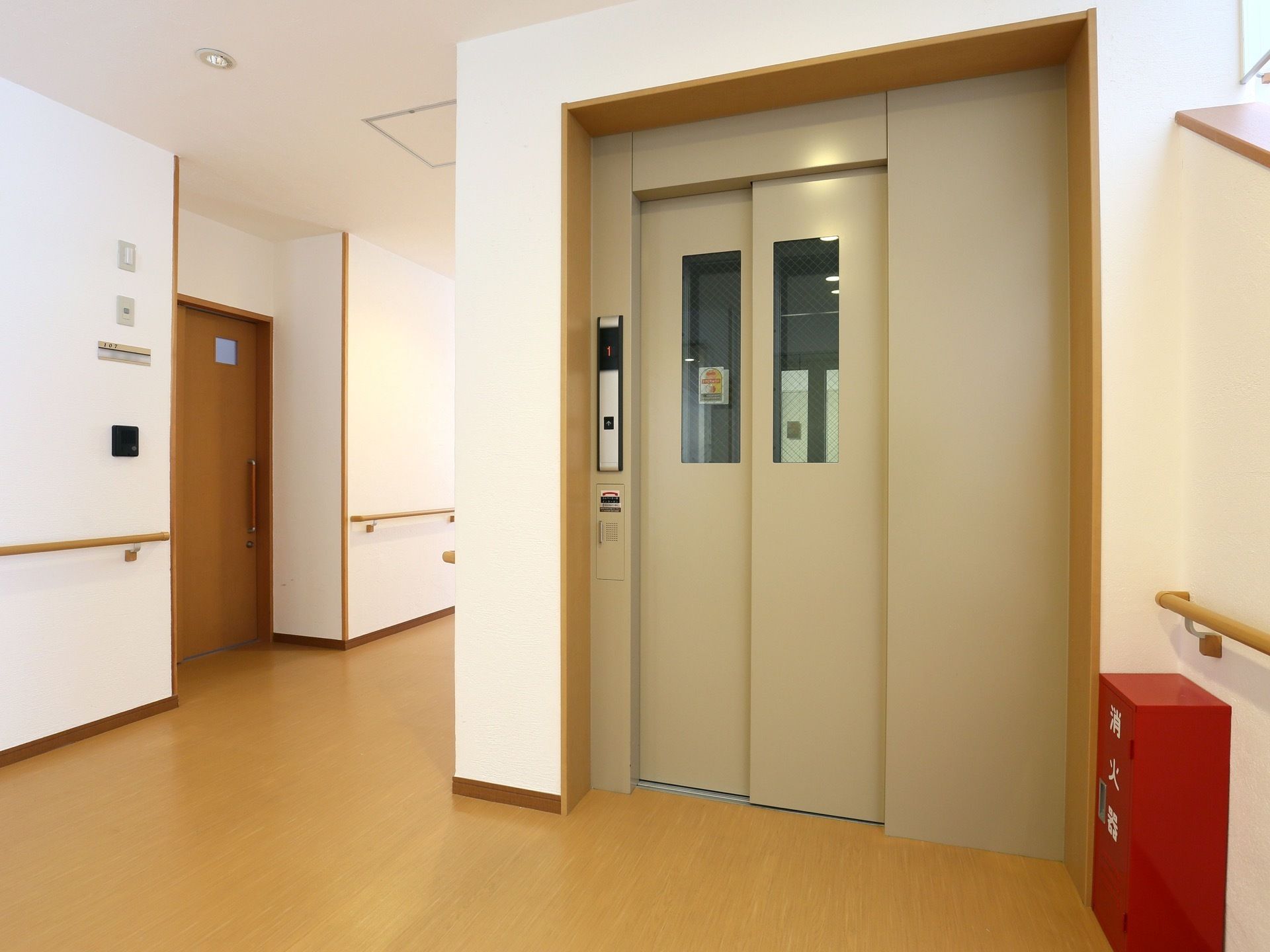クリーム色のエレベーターが茶色い廊下と白い壁に調和し、廊下に馴染んでいる。その扉には窓がついている。