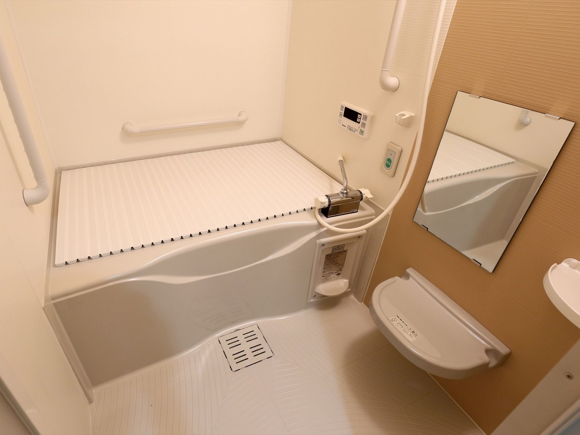 個浴用浴室である。奥に白い浴槽があり、蓋が閉められている。壁には給湯の操作パネルや非常ボタンが設置されている。