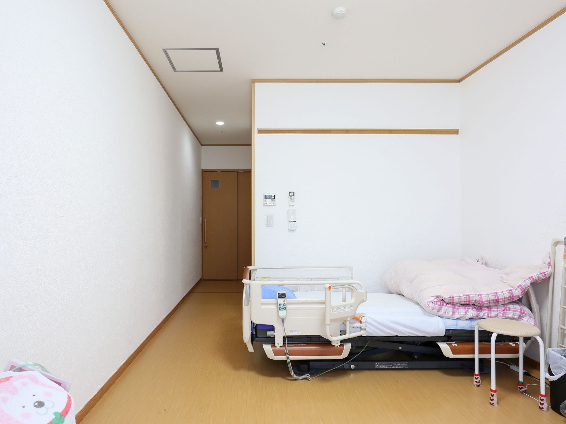 左奥に廊下へ出る引き戸扉がある。手前に居住スペースが広がり、布団の敷かれた介護用ベッドが壁際に置かれている。