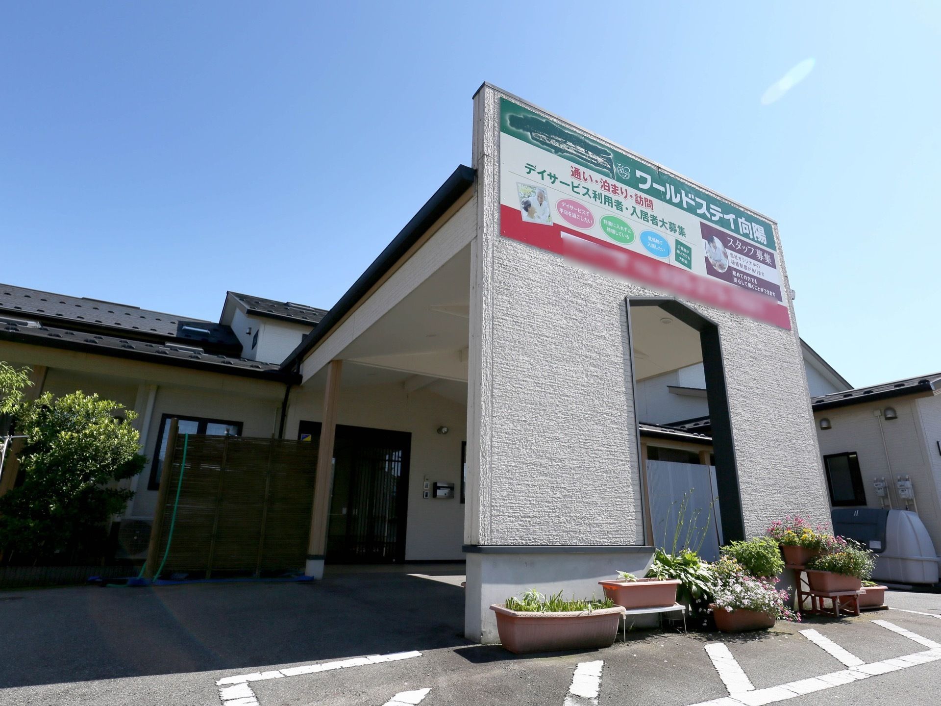 施設の入口前は屋根がせり出している。施設名が表示された大きな看板が取り付けられ、その下に鉢植えが飾られている。