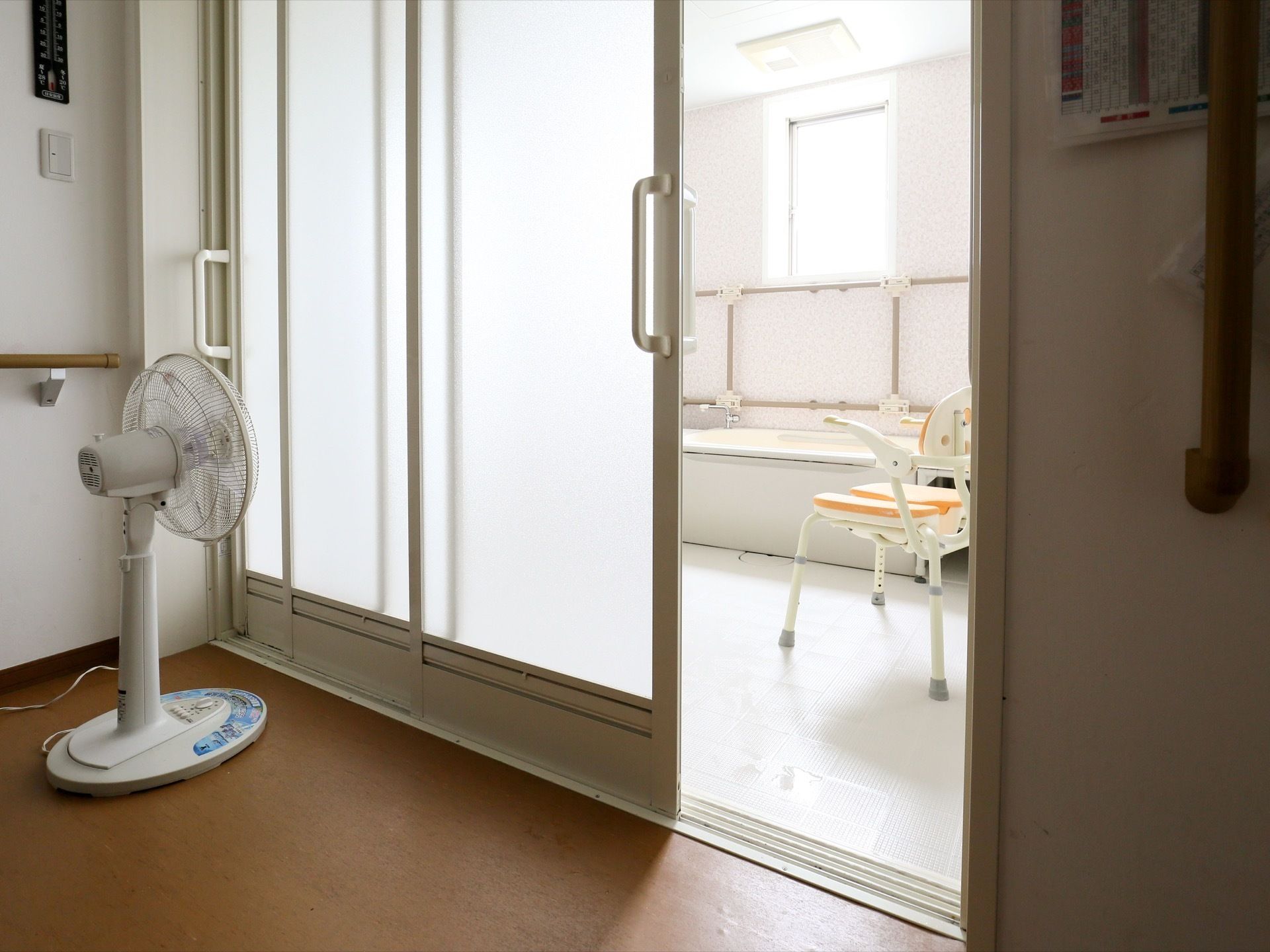 浴室の手前に脱衣室があり、左端に扇風機が置いてある。すぐ横の壁には、手すりや照明のスイッチが設置されている。
