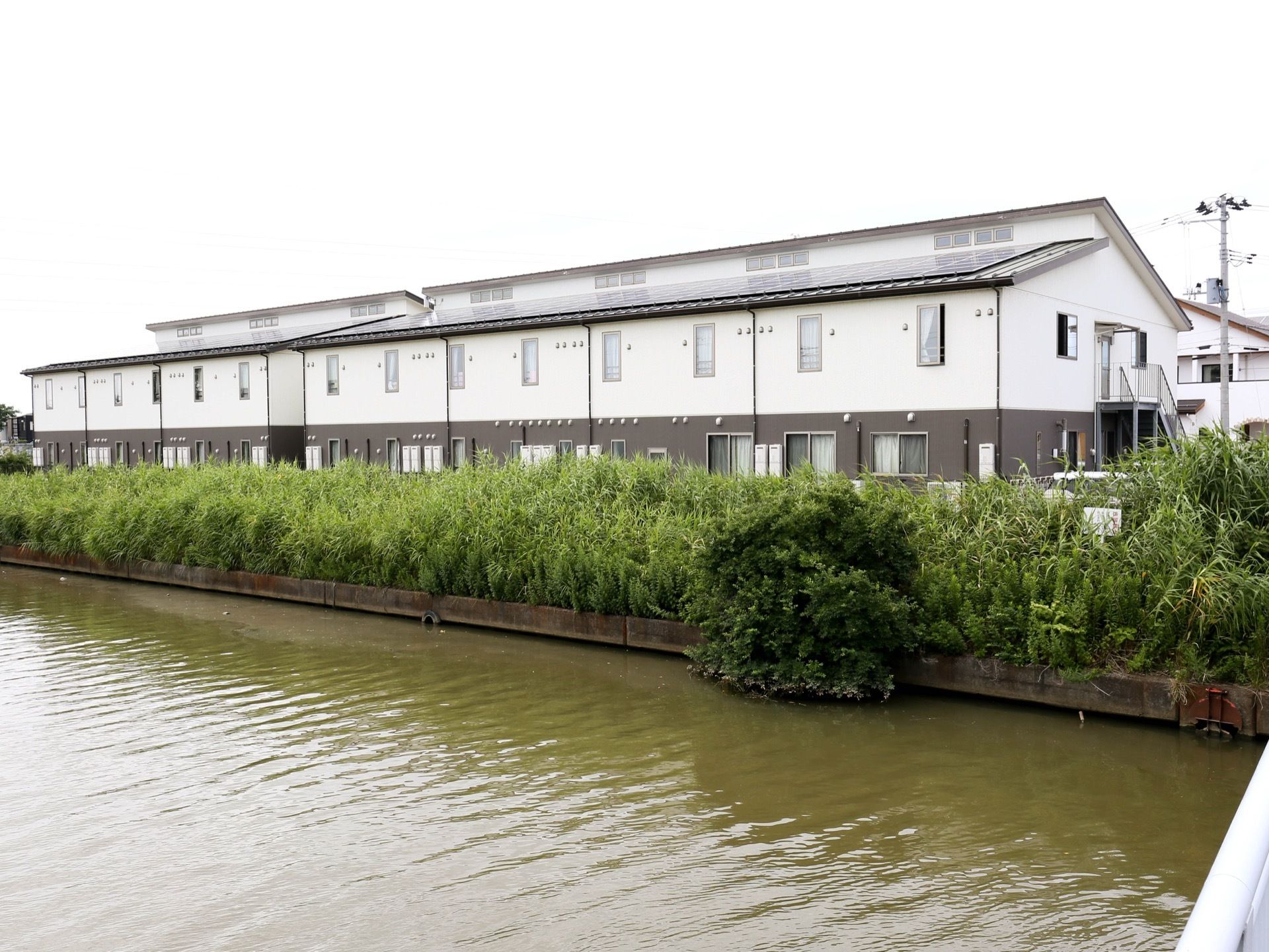 地上２階建ての施設の外観は、白と茶色のツートンカラーである。前方には川があり、その側には緑の草が生い茂っている。