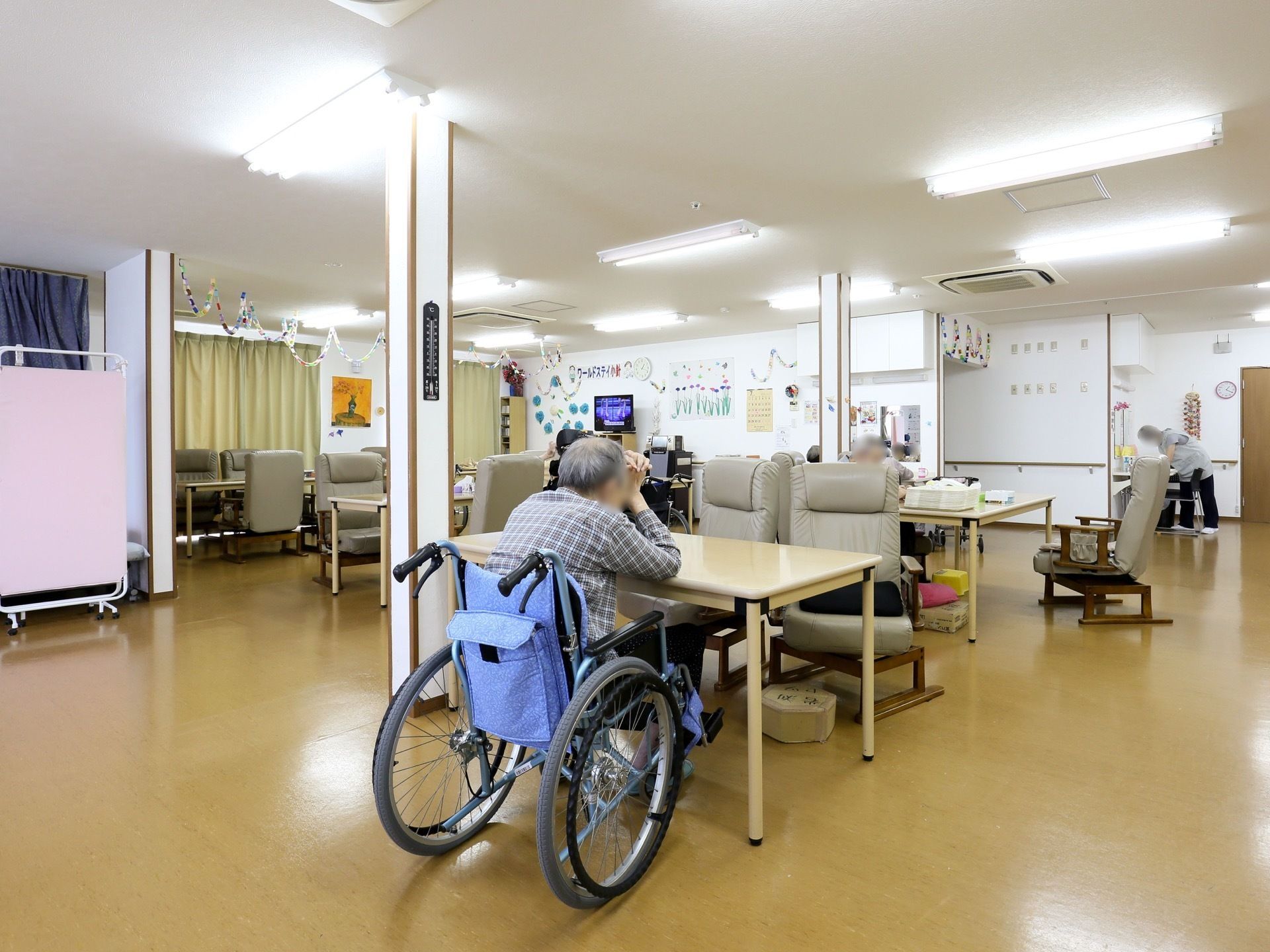食堂の奥にはテレビがあり、周囲の壁に色とりどりの装飾物が貼り付けられている。手前の柱に、黒い温度計が掛けてある。