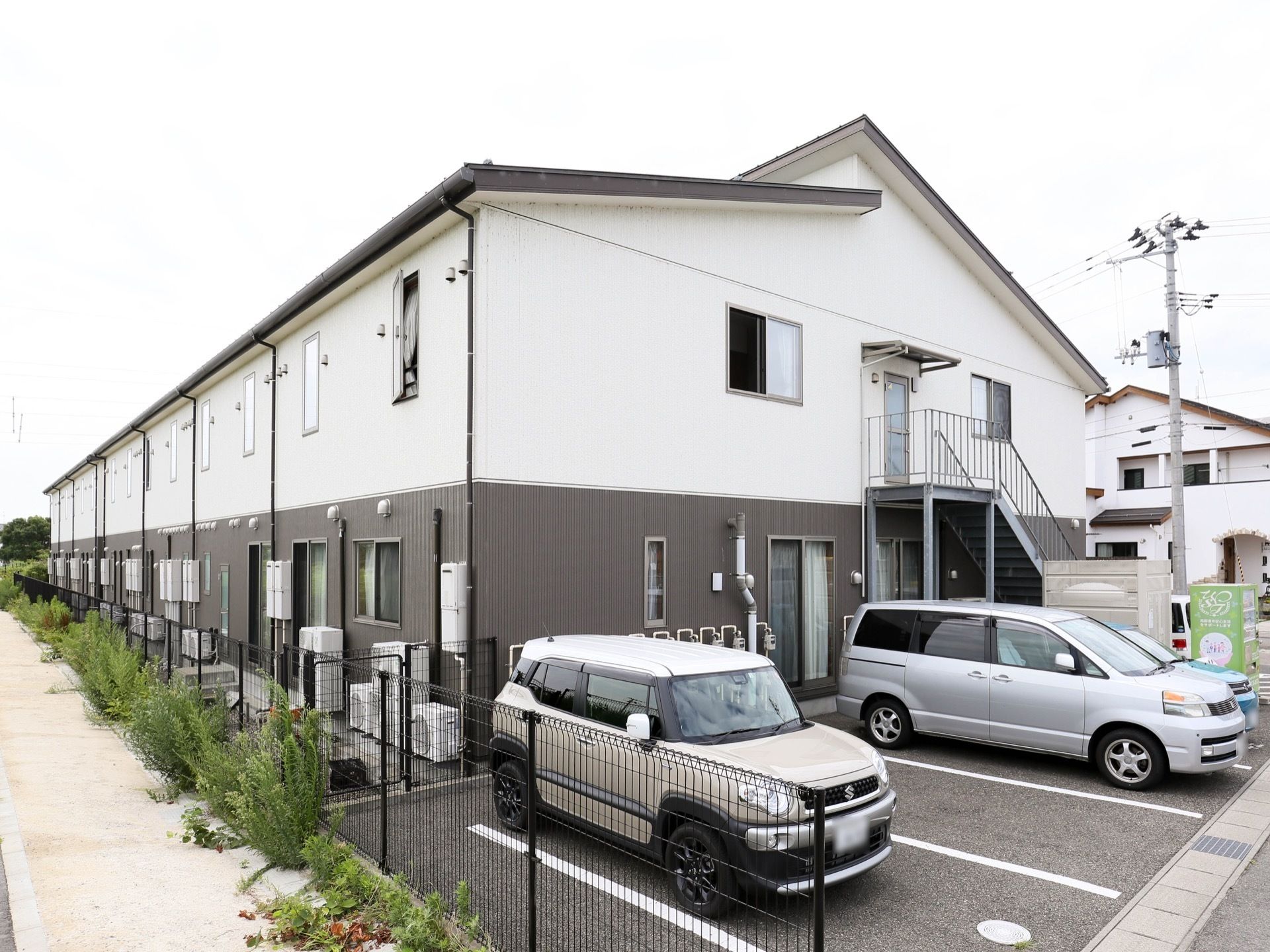 ライトグレーの非常階段が、外壁に取り付けられている。手前には駐車場があり、普通自動車やワゴン車などが並んでいる。