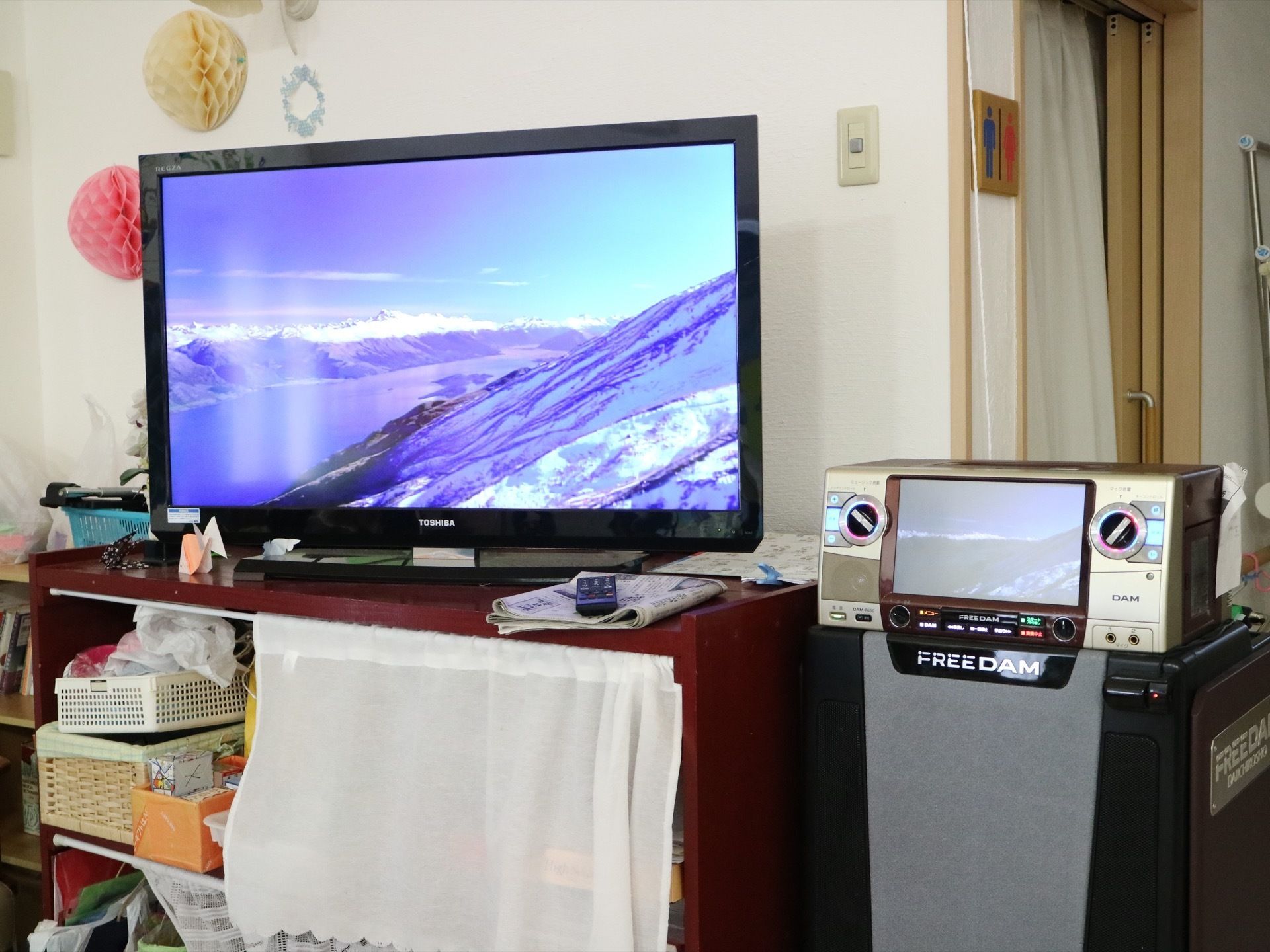 トイレ横の白い壁際には、収納棚の上にテレビが設置されモニター画面は点いている。横にはカラオケ機器が置かれている。