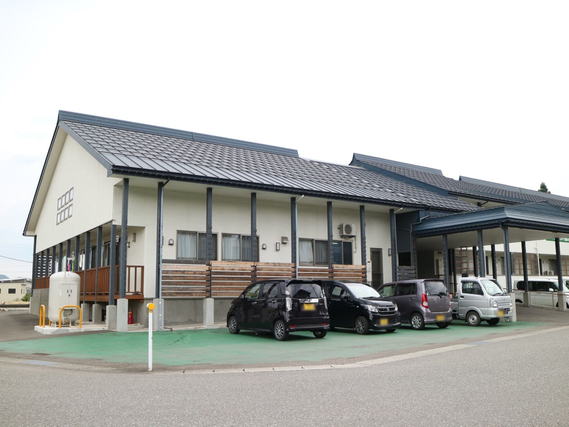 広く屋根付きのポーチが完備された平屋建ての施設に設けられた駐車場である。数台の乗用車が停車している。
