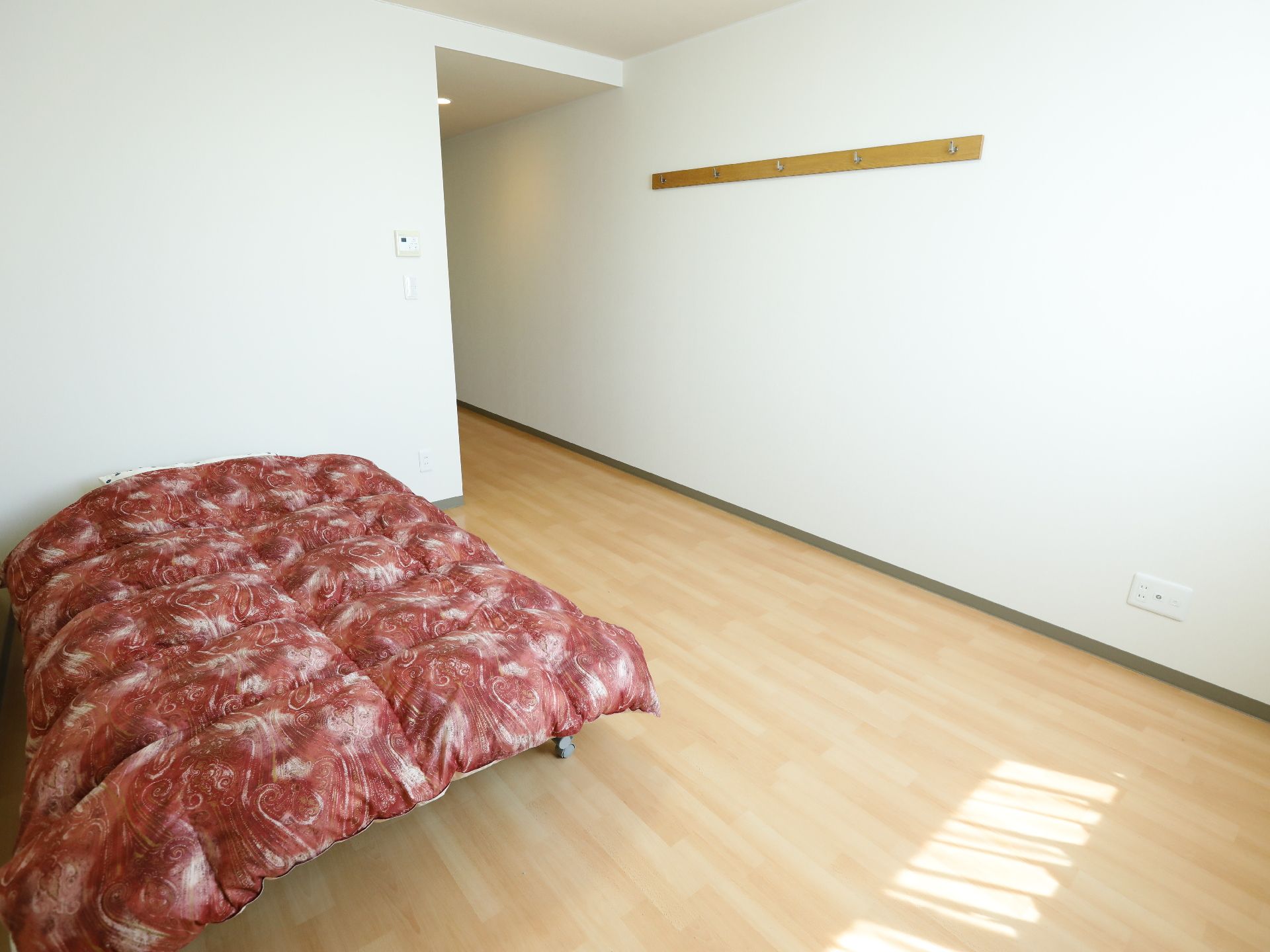 壁にフックがついている。ワンルームでフローリングの部屋には、ベッドが一台置いてある。赤い掛布団が見える。