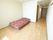 白い壁で、茶色のフローリングの部屋には、ベッドが一台置いてある。通路があり、キッチン、収納棚、扉が見える。