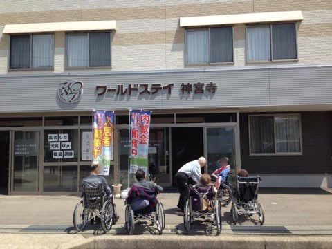 エントランス前の5名の車椅子を利用されている入居者様と、ホームの名前が書かれた外壁