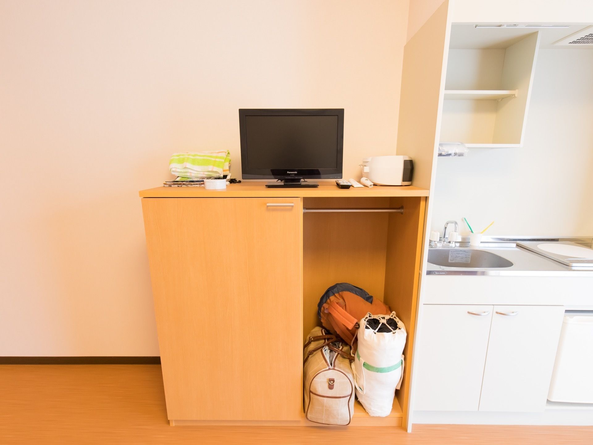 一口コンロのついたキッチンがある。シンク下に扉のついた収納と天吊りの収納が設置されている。茶色い収納棚の上に薄型テレビが置いてある。
