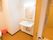 幅の広さがある洗面台が備わっている。大きい鏡と明るい照明が付いている。後ろにトイレがあり、掃除用具が置かれている。