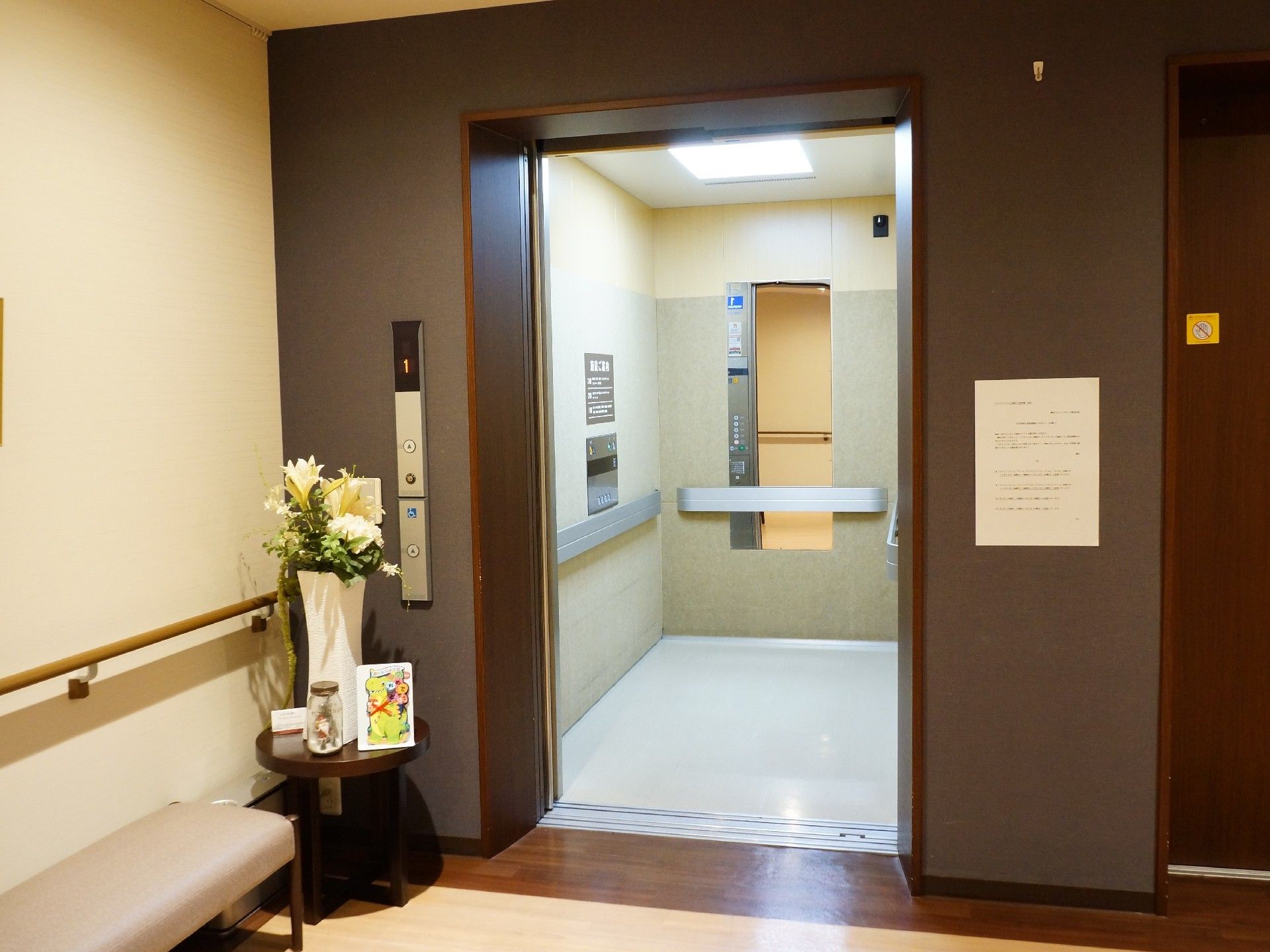 エレベーター前の壁とフロアはブラウンで、入り口横には花が飾られている。椅子が置かれ、壁には手すりが設置されている。
